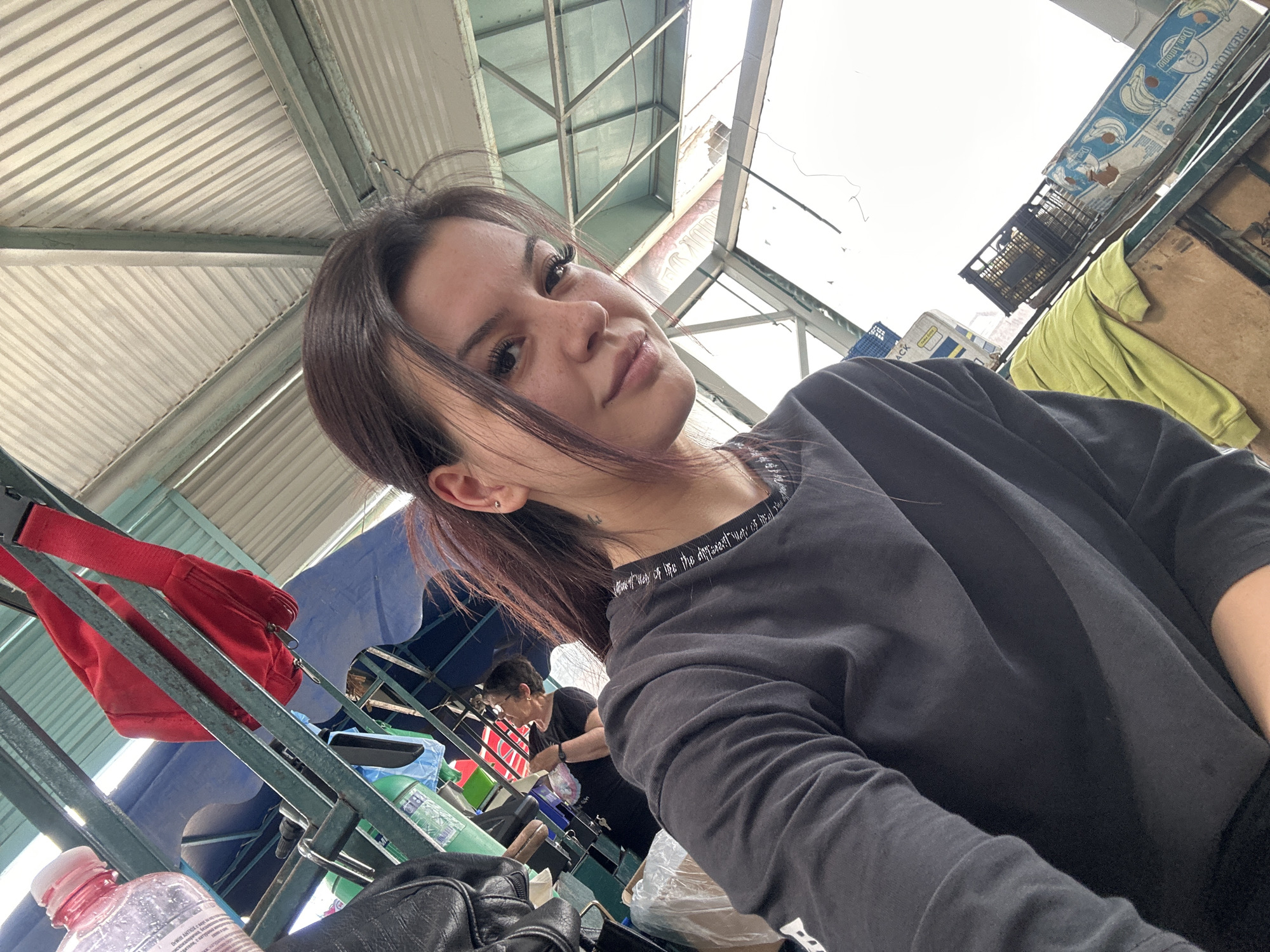A young woman is captured in a selfie, looking directly at the camera with a slight smile. She has dark hair, styled to the side, and is wearing a dark long-sleeved top with text on the collar that reads "the difference between the different ways of life." A small tattoo is visible behind her left ear.
The setting appears to be an indoor or covered market, characterized by a bright, corrugated metal roof and visible metal support beams overhead. The natural lighting suggests it is daytime.
In the background to the left, a red fanny pack or small bag hangs on a metal railing, with a clear plastic water bottle, partly obscured, visible below. Further back, a blue tarp or fabric is draped.
To the right, another person, an older woman with glasses and short hair, is visible in the mid-ground, engaged in an activity at what appears to be a market stall. Various items like plastic bags and containers are around her. Above her, a blue plastic crate, a black crate, and a cardboard box labeled "Don Antonio PREMIUM BANANAS" and "Product of Costa Rica" are stacked. A neon green garment is also hanging in this area. The overall scene depicts a lively market environment.