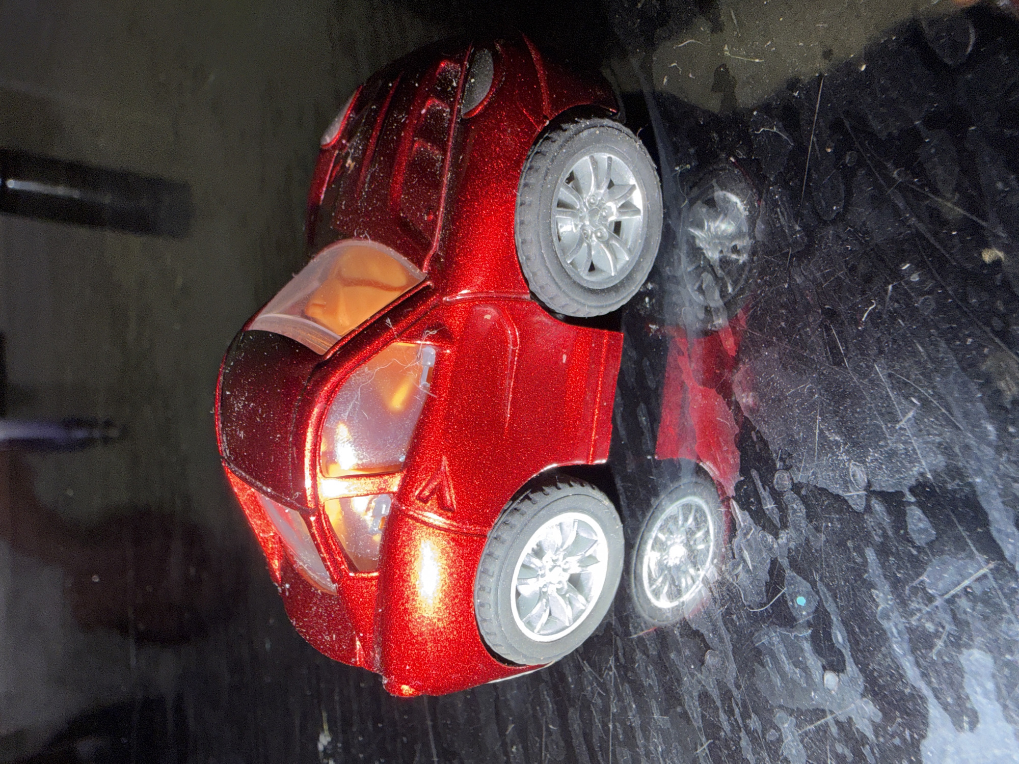 A shiny, metallic red toy car with large, exaggerated wheels sits on a dark, reflective surface. The car has a transparent orange interior and silver rims on its wheels. The surface it rests on appears to be a table or countertop, showing numerous scratches and some smudges, possibly water marks or dirt. In the background, out of focus, are blurry shapes that suggest a dimly lit indoor environment. No specific location in Bila Tserkva, Ukraine, can be identified from this image.