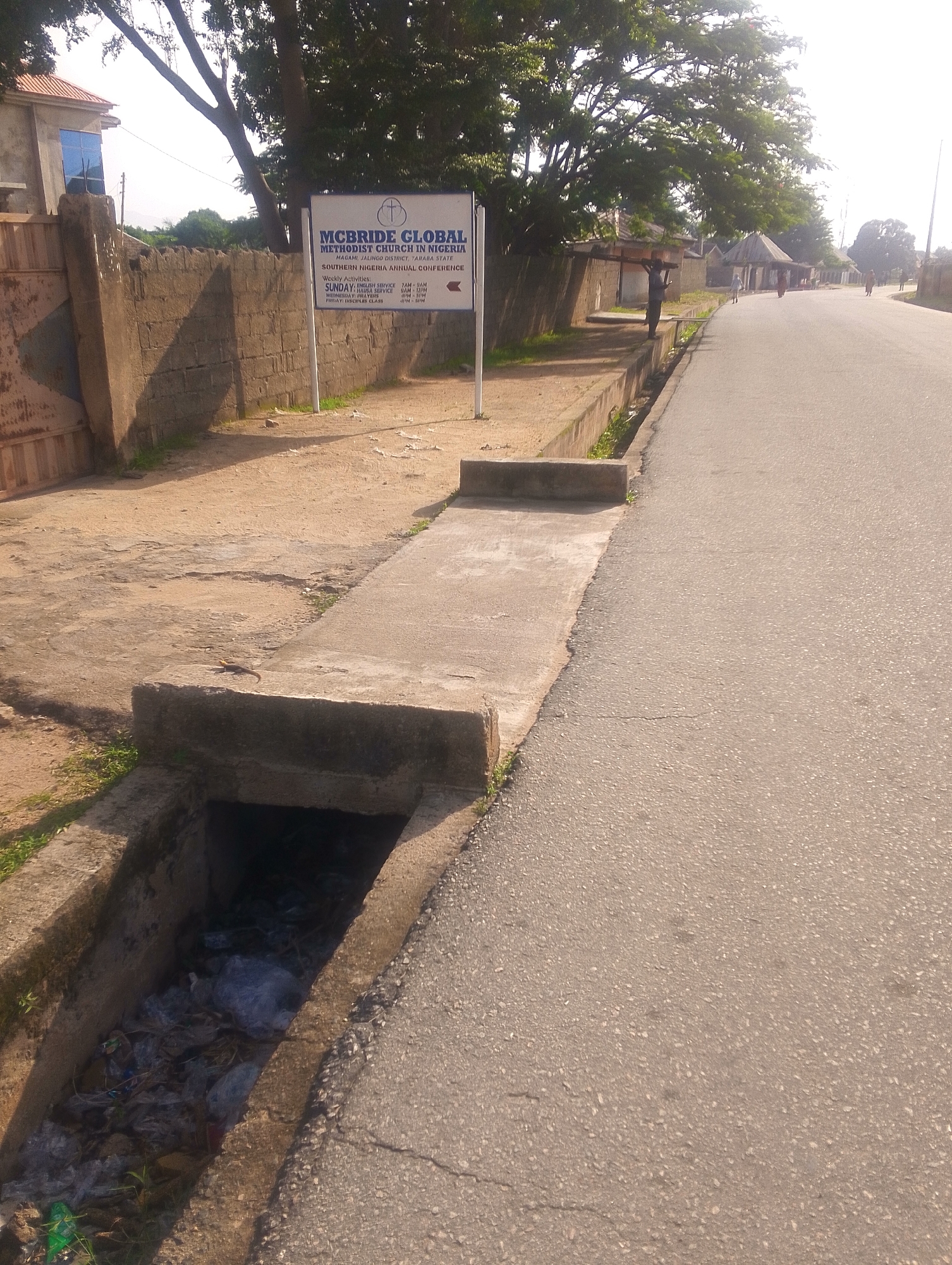 The image depicts a street scene in Jalingo, Nigeria. A sign in the foreground reads "McBRIDE GLOBAL METHODIST CHURCH IN NIGERIA" and provides details about the church's location and weekly activities, including Sunday service times and Wednesday prayer meetings. Next to the sign is a wall constructed from concrete blocks, and beyond that, a building with a red-tiled roof and blue windows. A large tree with spreading branches casts shadows on the scene.
To the right of the sign and wall, a paved road runs parallel to a concrete drainage ditch. The ditch appears to be filled with a significant amount of trash, including plastic bags and other debris. The road itself is asphalt, with some visible cracks and patches.
In the middle ground, several people are visible walking along the road or standing in the distance. One person is carrying a large wooden cross on their shoulders. Further down the road, there are more individuals and some small structures with thatched roofs. The overall setting suggests a residential or community area.
The lighting indicates it is daytime, likely late morning or early afternoon, with the sun casting distinct shadows. The weather appears to be clear. The scene conveys a sense of everyday life in a Nigerian town, with the presence of religious institutions, a busy road, and the common issue of waste management reflected in the littered drainage.