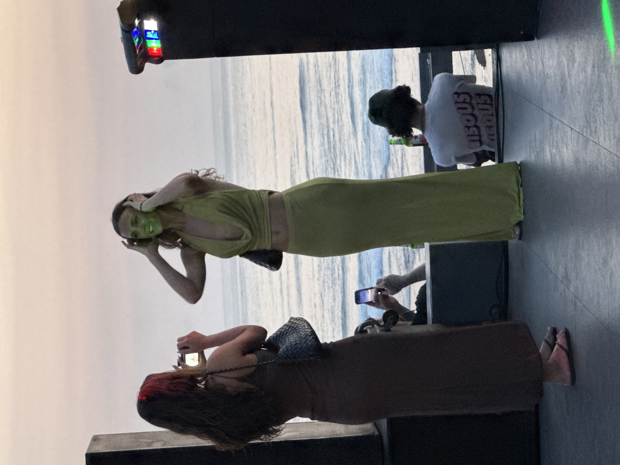 The image, taken in Kuta Selatan, Indonesia, depicts a terrace overlooking the ocean at dusk. Three women and one person wearing a t-shirt are visible. One woman, wearing a long, green dress and with green-toned skin, is holding her hands up to her head, seemingly posing. Another woman, with long, dark hair with red highlights, is taking a photo of her with a mobile phone. A third woman, wearing a long brown dress, is standing nearby. Another person is seated at a table in the background, wearing a white T-shirt with bold print and a green cap, and holding a bottle. The ocean is visible in the background, appearing calm. Another person is holding a phone as well. The setting includes black structures, possibly part of the terrace or furniture, and there's some lighting equipment with colorful lights.