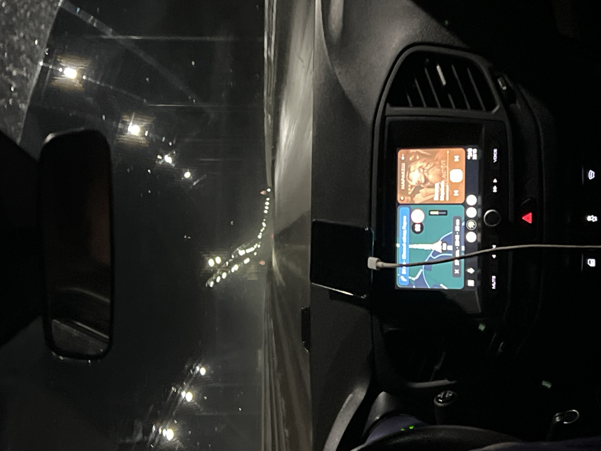 The image captures the interior of a car at night, viewed from the driver's perspective looking out through a wet windshield. The car appears to be in Zvyozdny, Russia.
A prominent car infotainment system dominates the right side of the frame, displaying a split screen interface. The upper half shows a music player with an album cover featuring a person, along with the text "МАРАКЕШ" and "nora." The lower half of the screen displays a navigation map, illustrating a blue route with estimated travel times, such as "12 min" and "20 min." A white charging cable is plugged into the bottom of the infotainment unit.
Around the screen, elements of the car's dashboard are visible, including an air vent above and several buttons to the right, one of which is a red triangular hazard light button. Other visible text includes "VOICE" and "MUTE" near the screen, and a digital clock showing "0:23." A partial view of the steering wheel is at the bottom right.
Through the windshield, the dark ambiance of a night drive is evident. Numerous bright, scattered points of light, presumably streetlights, are reflected and streaked across the glass, indicating that the windshield is wet, suggesting rain or recent precipitation. A portion of the driver's side rearview mirror is visible on the far left, reflecting the dark surroundings. The scene depicts activity of driving at night, with active navigation and music playback.