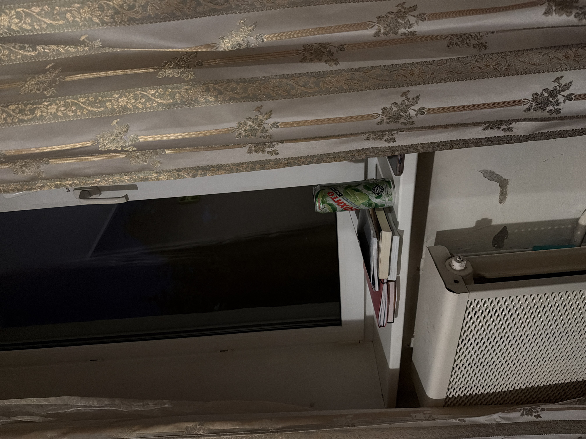 The image shows a close-up view of a windowsill in an indoor setting. On the windowsill, there are several objects: a stack of books, a can of what appears to be a carbonated beverage with a label featuring green and white text, and a smartphone. The windowsill is situated in front of a window with curtains drawn to the right side of the frame. The curtains are decorative, featuring a pattern of vertical stripes and floral motifs in gold and beige. To the left of the windowsill, a portion of a white radiator with a patterned grille is visible. The overall lighting suggests it may be nighttime or a dimly lit room. The background of the window is dark, indicating it is either night or the outside is not brightly illuminated. The wall beneath the windowsill has some minor imperfections. The scene is static and devoid of human presence or activity.