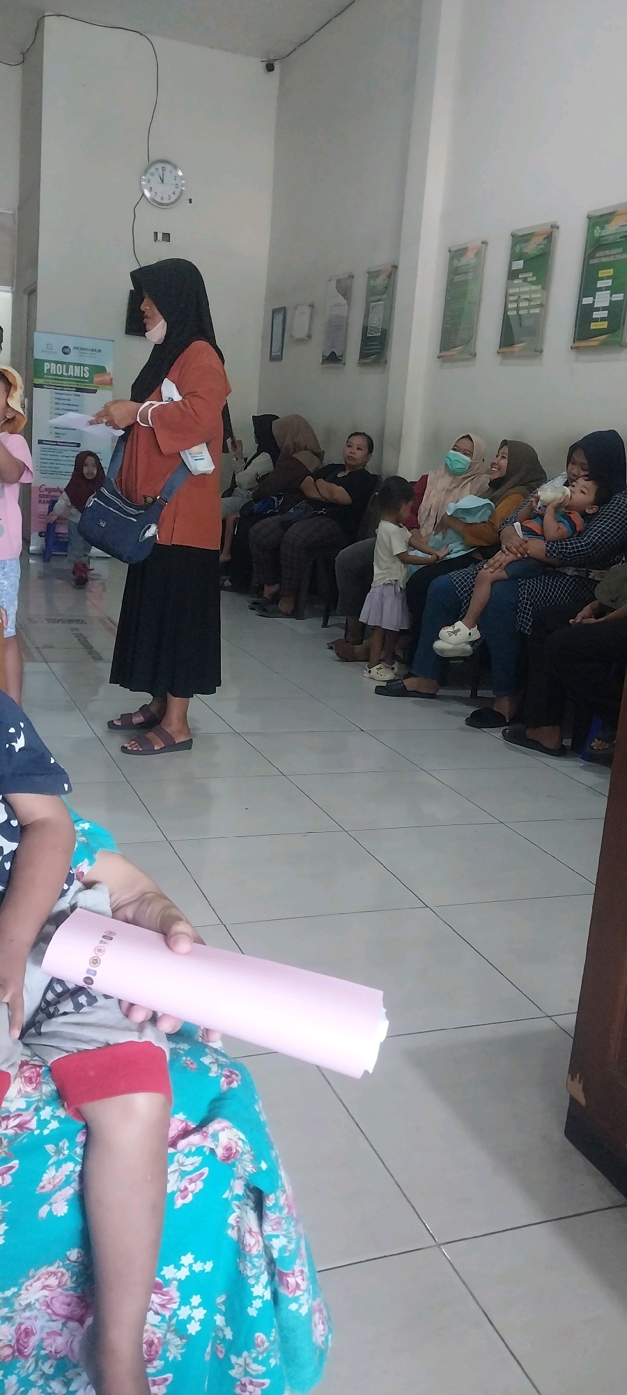 The image depicts a waiting area, likely a clinic or hospital, in Kalijaten, Indonesia. Several individuals, predominantly women and children, are seated on a bench along the wall, awaiting service. A woman wearing a black hijab and an orange top stands to the left, holding a paper and a blue bag. A child in a pink outfit is partially visible to her left. On the right, a child is being fed from a bottle by a woman. Another child in a light purple skirt is standing. Posters are displayed on the wall. The floor is tiled.