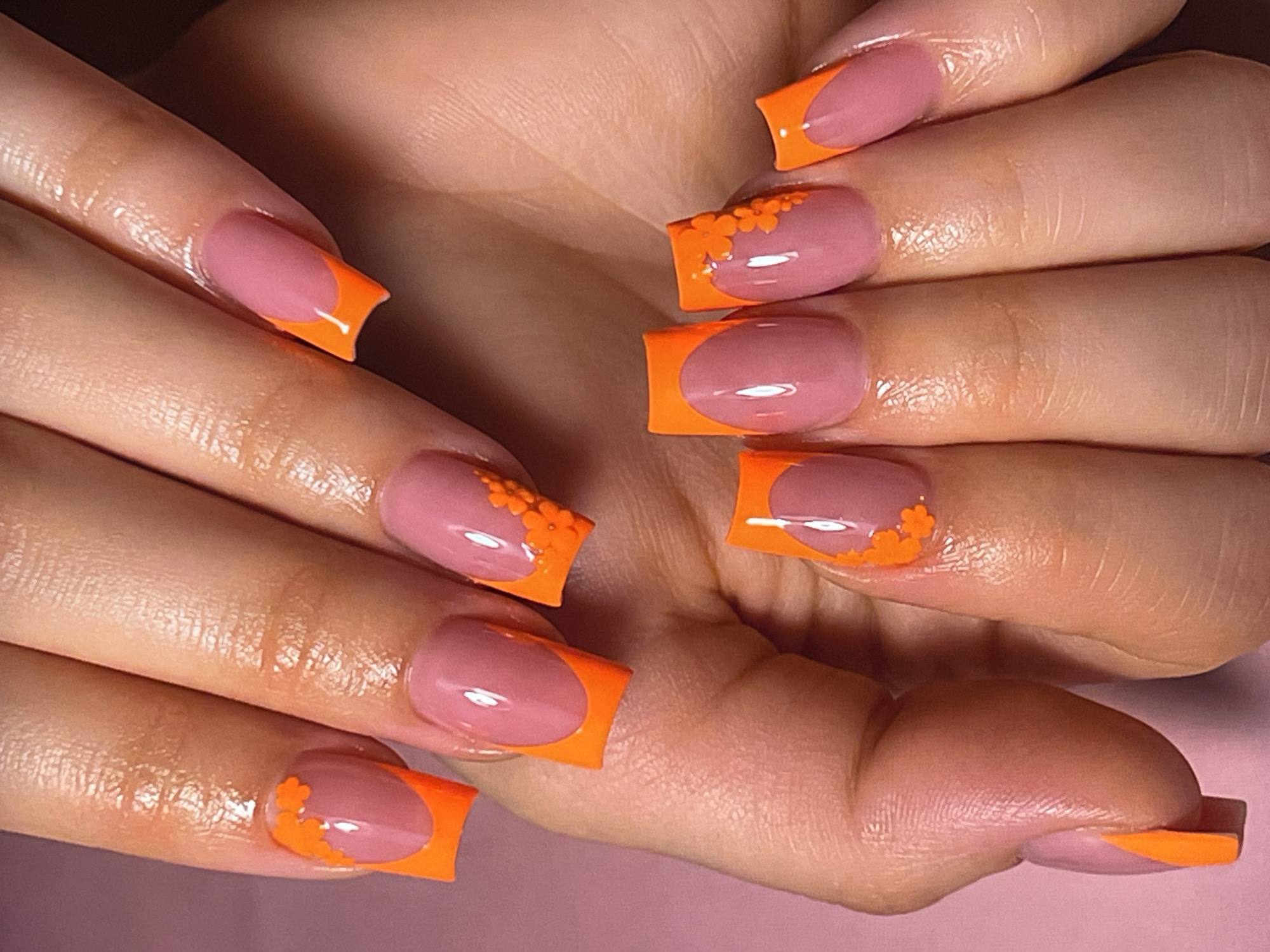 An overhead close-up shot shows a person's hands with a manicure. The nails are long and square with rounded edges, and they have a pink base color. The tips of the nails are painted with a bright orange French tip, and some of the nails have small orange floral accents. The hands are positioned against a light pink background, and the lighting creates subtle shadows that highlight the texture of the skin and the glossy finish of the nail polish.