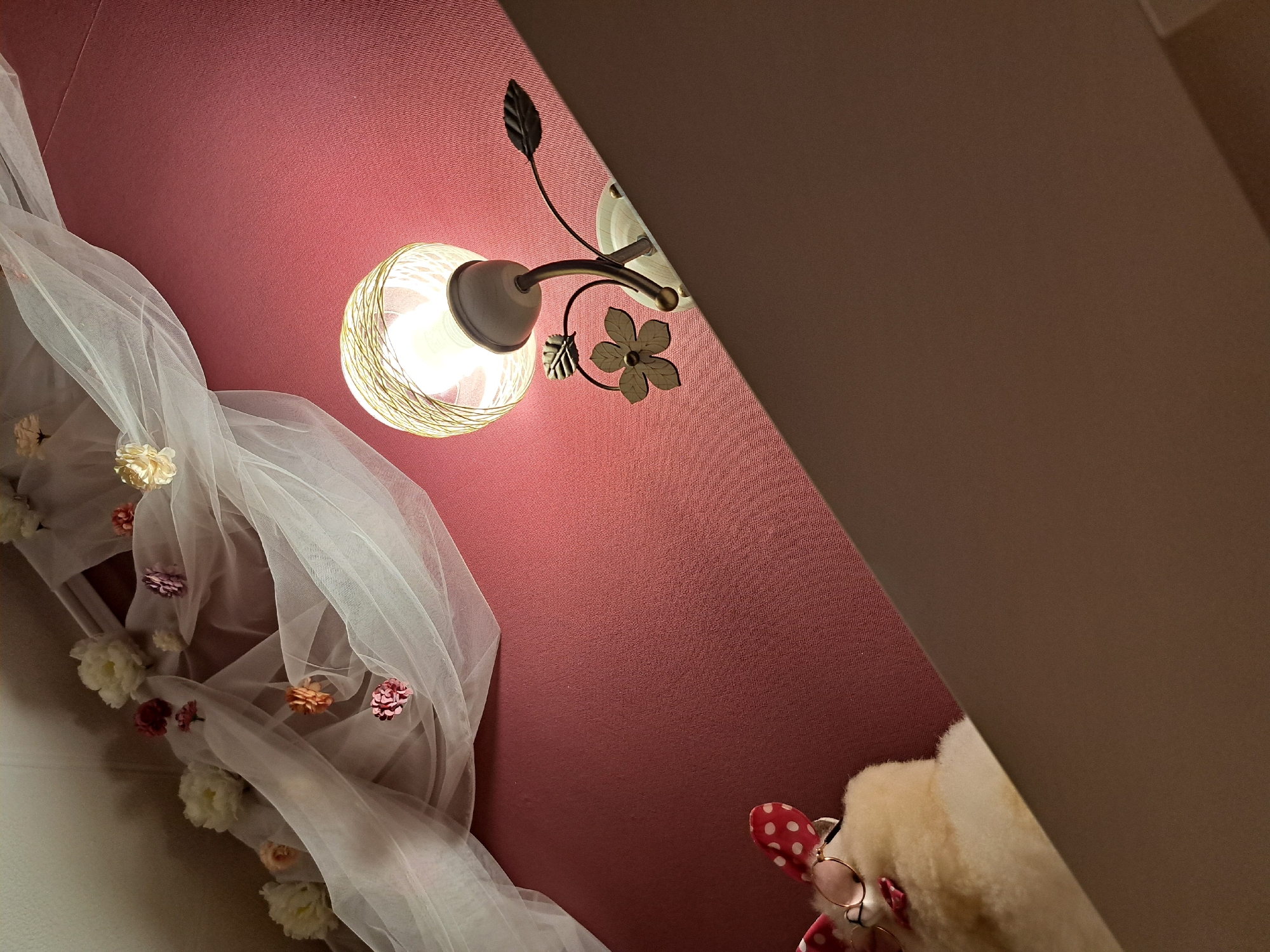 This image captures an indoor scene from a low, diagonal perspective, looking upwards at a textured, reddish-pink wall. A decorative wall-mounted lamp, featuring an antique-style metal arm with leaf and flower embellishments, is prominently lit, casting a warm glow. Its shade is a clear glass globe wrapped with thin, light-colored wires.
To the left and bottom of the frame, a sheer, white fabric, resembling tulle or organza, is draped elegantly. This fabric is adorned with several small, artificial flowers in cream, pink, and orange hues. In the bottom right corner, the upper part of a cream-colored plush toy is visible. The toy wears round, gold-rimmed spectacles and has a red, polka-dotted ear or bow. A flat, dark brown surface cuts across the top right portion of the image, creating a sharp diagonal line.
The warm, artificial light from the lamp suggests an indoor setting, possibly during the evening or night, contributing to a cozy and whimsical ambiance. No people are present, and no specific activity is taking place. There is no visible text within the frame.