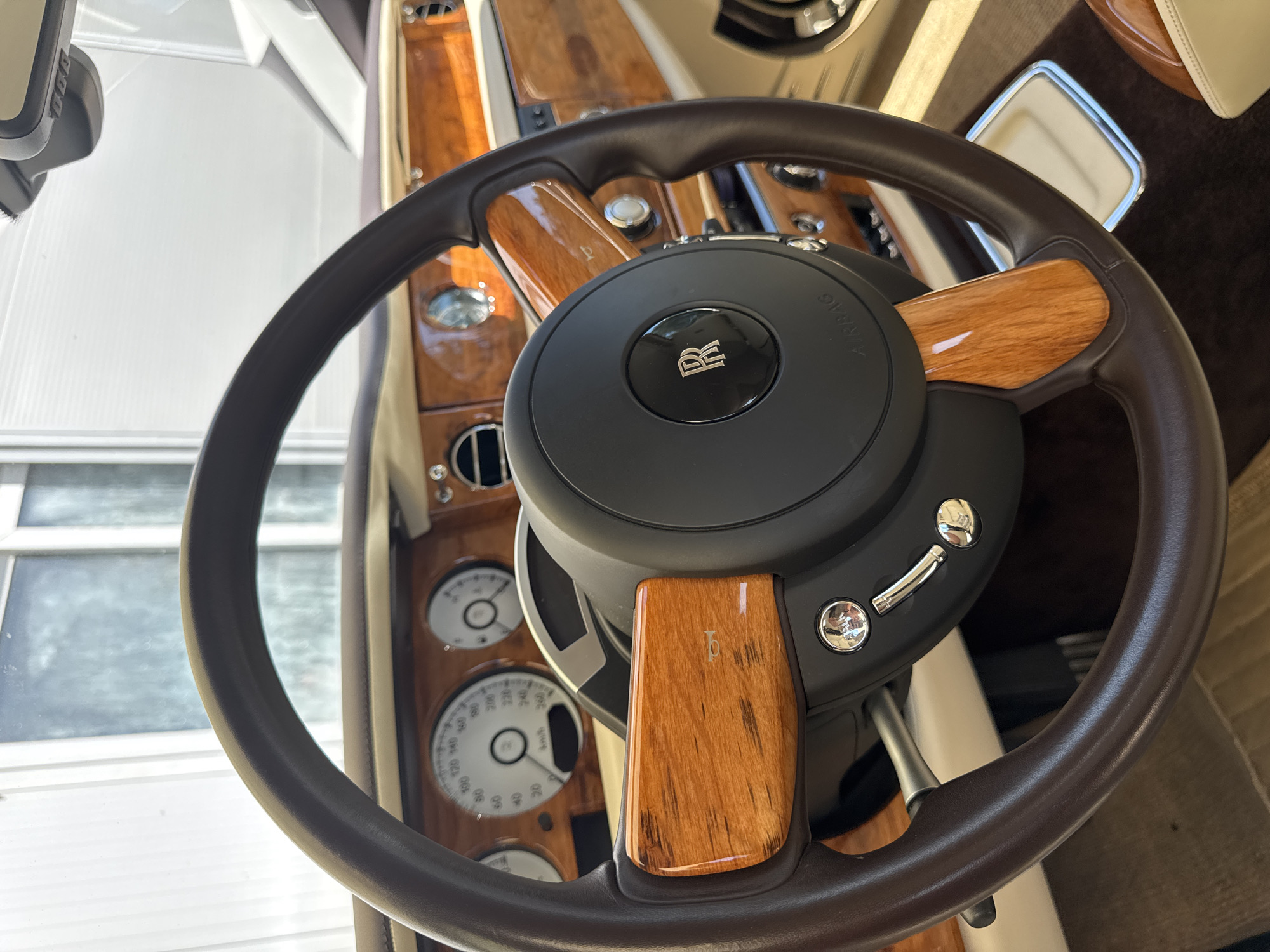 The image shows a close-up view of the interior of a luxury car, likely a Rolls-Royce, with a focus on the steering wheel and dashboard. The steering wheel is upholstered in dark brown leather and features polished wood accents. The center of the steering wheel displays the Rolls-Royce "R" logo on a black circular emblem, with "AIRBAG" text below it. To the left of the steering wheel, a speedometer is visible, with markings in kilometers per hour. The dashboard is adorned with rich, glossy wood trim. Chrome buttons and levers are also present on the steering wheel and the dashboard. The car appears to be parked indoors, possibly in a showroom or a garage, as suggested by the background which shows large windows and a bright, diffused light. There are no people visible in the image. The scene suggests a static, well-maintained environment, characteristic of a luxury vehicle display. The location is identified as Dub, Montenegro.