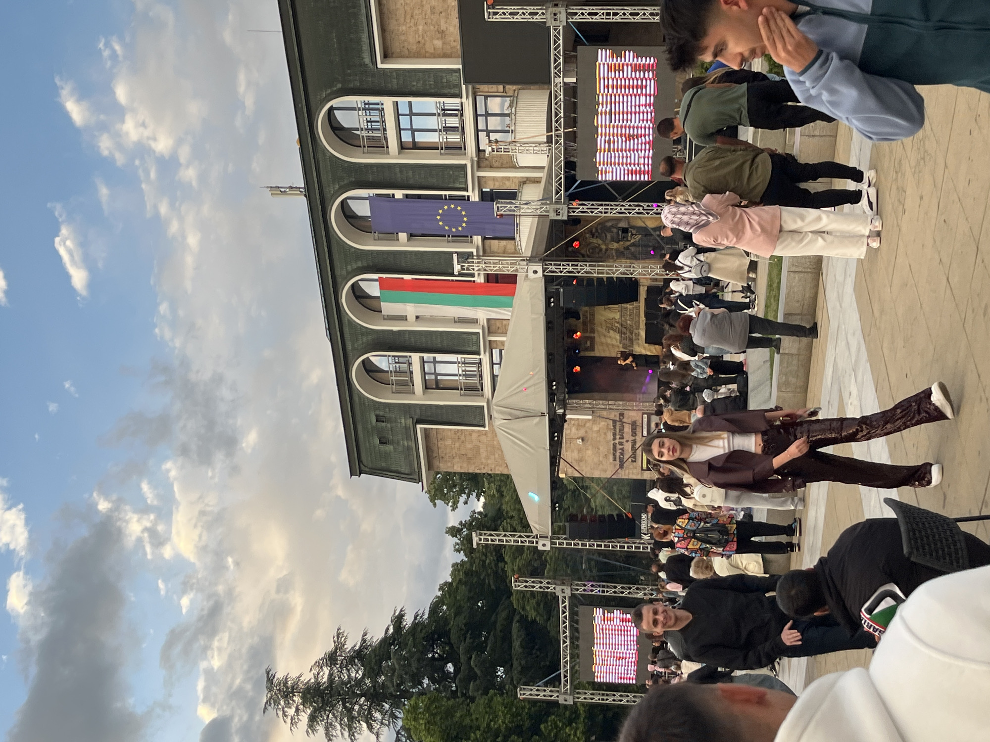 The image shows an outdoor event in front of a building with a green roof and three arched windows. The flags of Bulgaria and the European Union are displayed. A stage is set up with scaffolding and screens, and a crowd of people is gathered. A woman in a brown blazer and pants stands out in the foreground. Trees are visible in the background. The location is Unknown, Unknown.