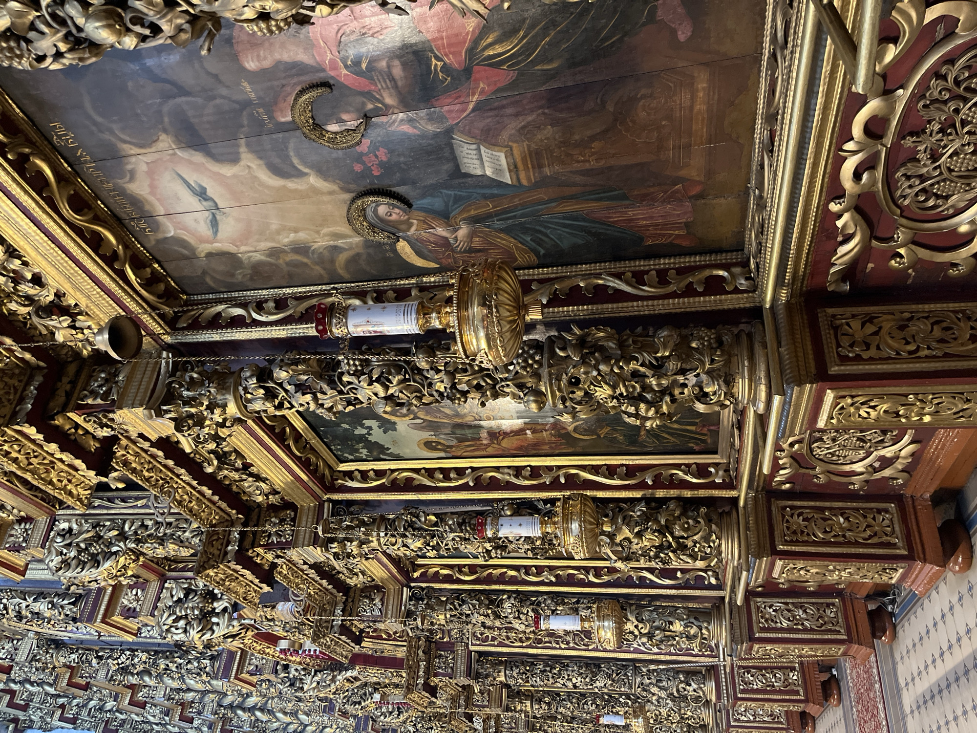 The image depicts the interior of the Trinity Cathedral in Pskov, Russia, featuring a richly decorated iconostasis. The iconostasis is adorned with intricate gold-leaf ornamentation and features several large icons. Hanging from chains are ornate golden candle holders, each holding a white candle with red detailing at the top. The walls are covered in dense, detailed carvings and gilded elements. The floor is tiled with a patterned design. The lighting is dim, casting shadows and highlighting the ornate details of the cathedral's interior.