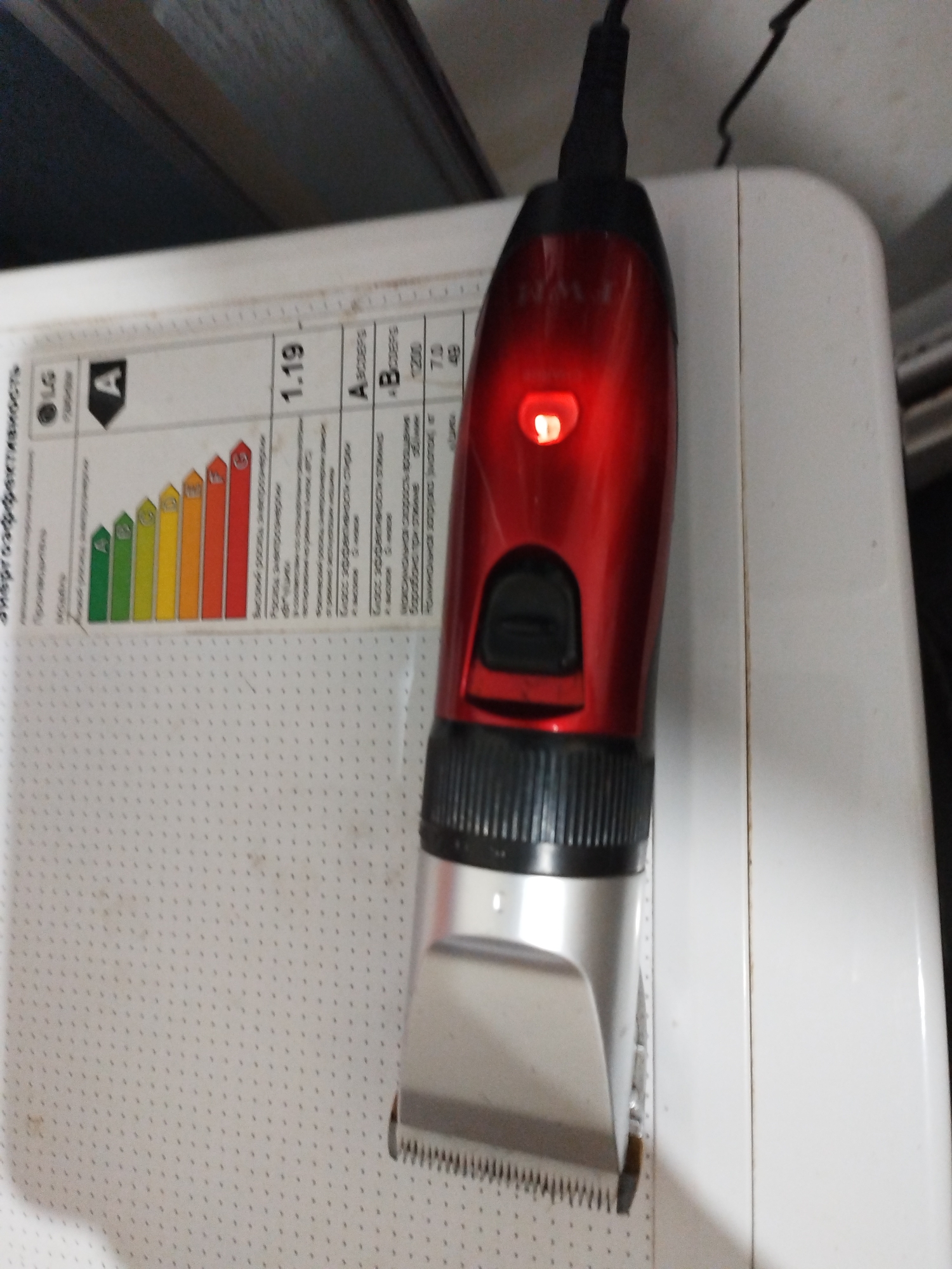 A red and silver electric hair clipper is charging on a white appliance, possibly a washing machine, with an energy efficiency label visible. The clipper has a red indicator light illuminated. The appliance has a textured surface with a grid pattern. The background appears to be indoors, with a dark surface above the appliance. The scene is photographed from a slightly elevated angle, focusing on the clipper and the appliance.