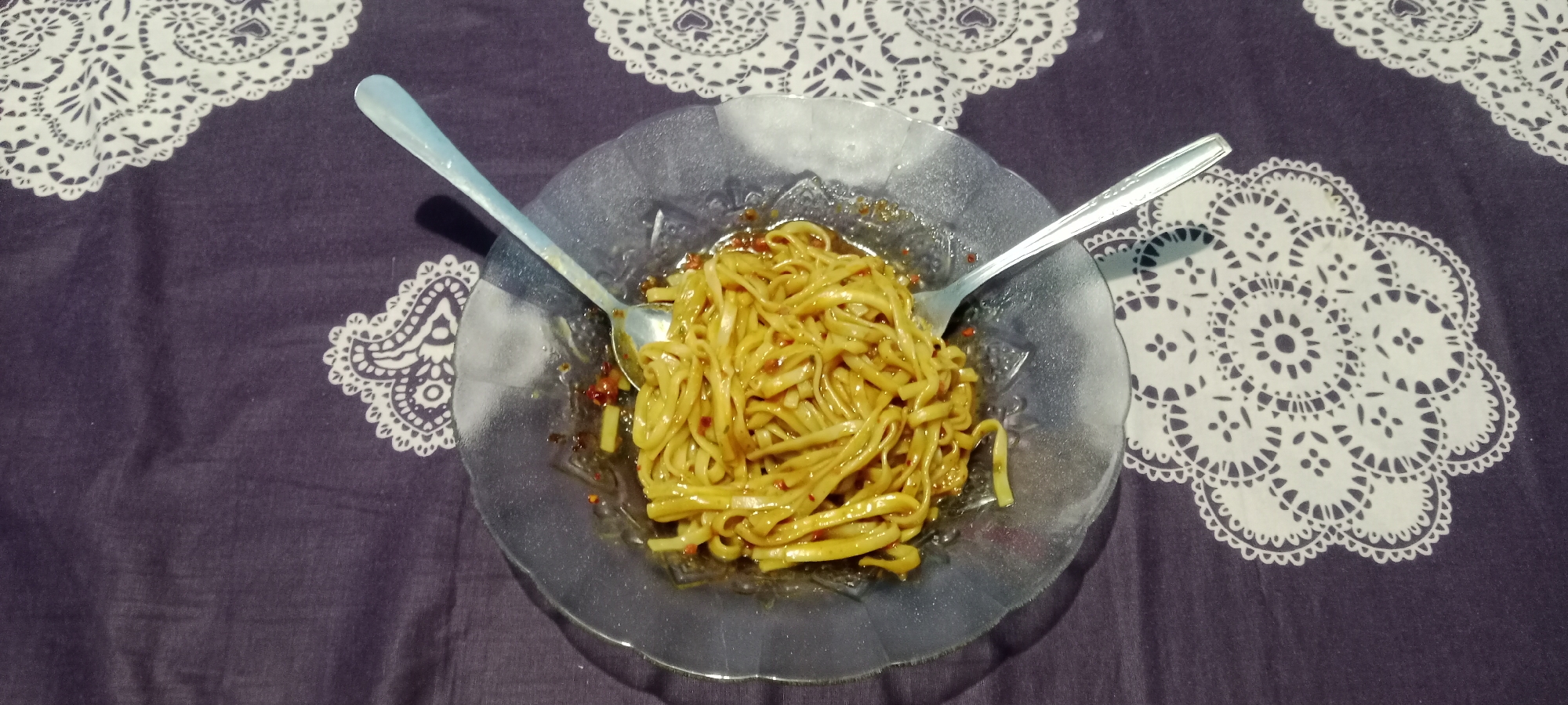 A plate of noodles is displayed on a dark purple tablecloth with a white lace pattern. The plate is transparent glass, and it is filled with noodles coated in a reddish-brown sauce. Two metal spoons are placed on either side of the plate, with one spoon partially submerged in the noodles. The lighting suggests an indoor setting, possibly a home. The image does not contain information to determine its location in Suka Tani, Indonesia.
