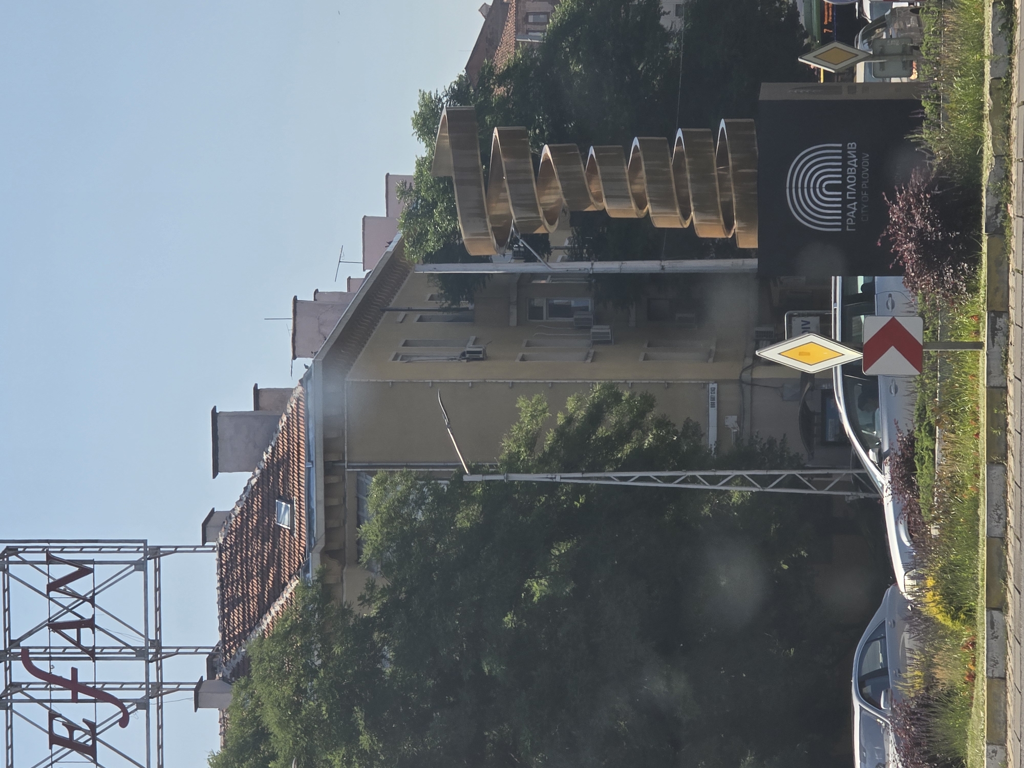 The image captures a view of a city street in Plovdiv, Bulgaria, featuring a modern spiral sculpture. The sculpture is gold-colored and twists upwards, standing on a black square base that displays the text "ГРАД ПЛОВДИВ" and "CITY OF PLOVDIV" along with a logo of concentric arches. To the left of the sculpture, a light pole with a bent lamp extends across the frame. Behind the sculpture and light pole, a building with a pale yellow facade and numerous windows is visible. The building has a tiled roof with several chimneys. Adjacent to the building, a large evergreen tree with dense foliage partially obscures the view. Further in the background, more buildings with tiled roofs and chimneys are seen, along with a bright blue sky. In the foreground, two silver cars are parked on a street, and a median strip with low-lying bushes and flowering plants separates the roadway from the sidewalk. Road signs, including a diamond-shaped yellow sign and a red and white directional sign, are positioned on the median. A large billboard with red lettering "EFAN" is visible at the top of the image, mounted on a metal frame. The scene appears to be a sunny day with clear weather conditions.
