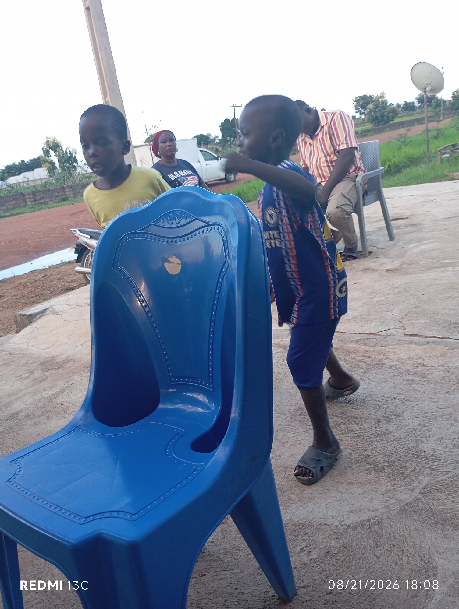 A full shot image shows a young boy in a blue and red striped shirt and blue shorts walking past a large blue plastic chair. The boy is wearing grey sandals. In the background, another young boy in a yellow shirt looks on, and a woman wearing a red headscarf and a black t-shirt stands behind him. Further in the background, a man in a striped shirt and khaki pants sits in a gray plastic chair. A white pickup truck is parked behind the woman, and trees and a satellite dish are visible in the distance. The scene appears to be outdoors, on a concrete surface. The image is dated 08/21/2026 18:08.