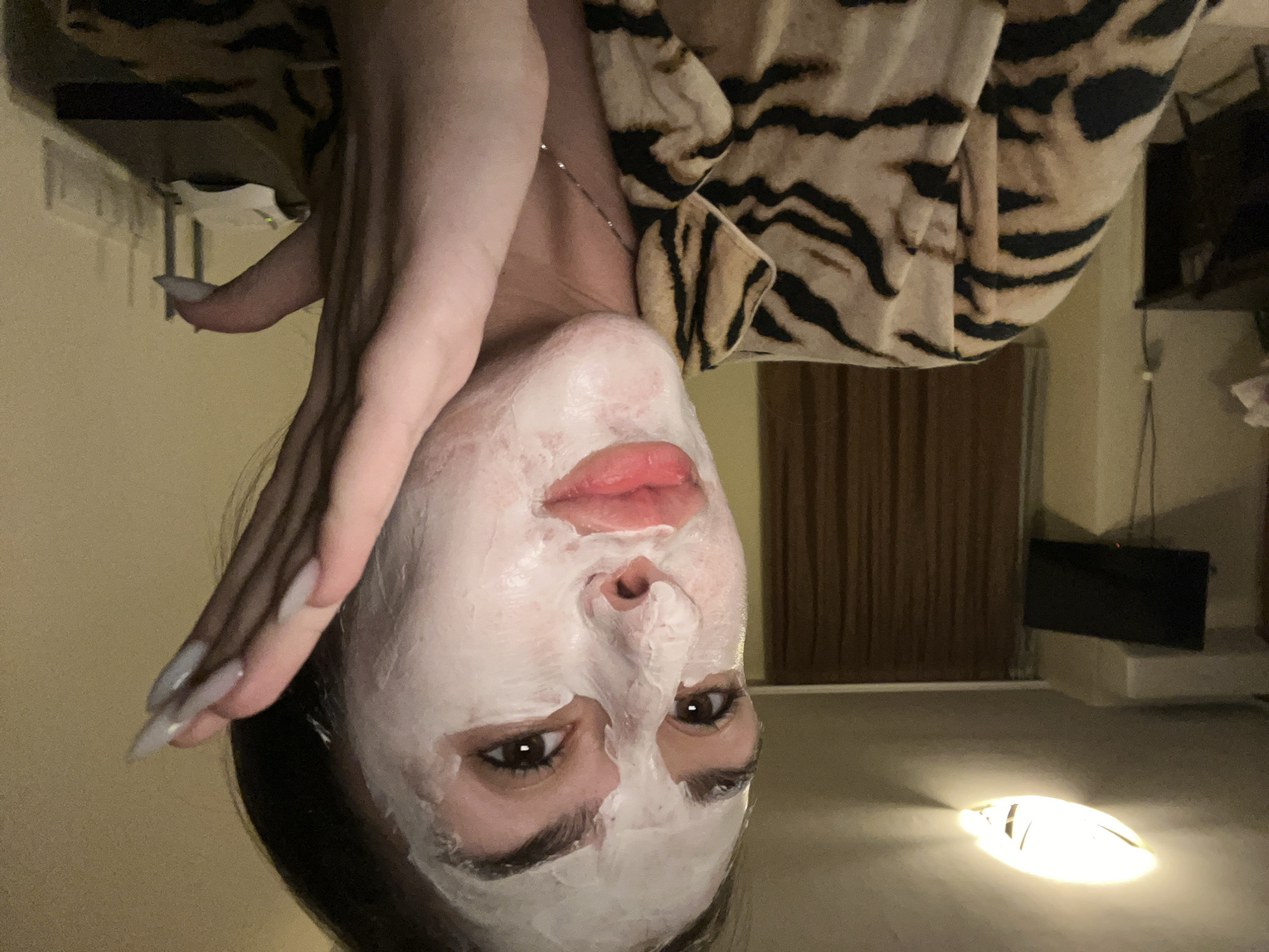 A female with fair skin and a white facial mask is seen in a close-up, upside-down view. Her right hand is positioned near her face. She has long, pointed nails. The woman is wearing a tiger-striped robe or top. A thin necklace is visible on her neck. Behind her is a dimly lit interior. The background includes a dark, rectangular object mounted on a wall, likely a television. A light fixture on the floor is also visible. The location appears to be indoors, possibly a residence. There is no information available to determine that this image was taken in Sofia, Bulgaria.