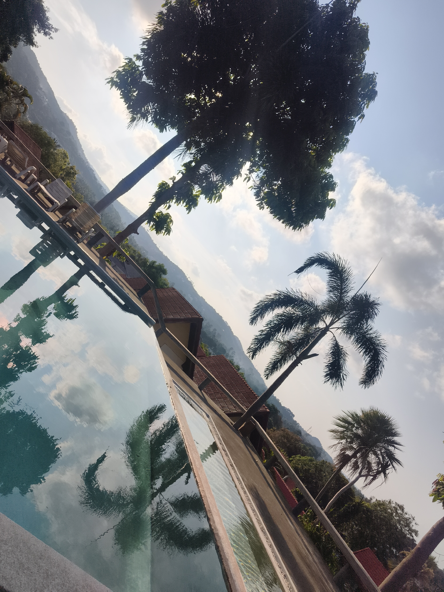 This image captures a serene scene in บ้านบางรักษ์, Thailand, featuring a swimming pool and surrounding landscape. The pool's surface reflects the sky and nearby trees, creating a mirror-like effect. Visible are lounge chairs on a deck adjacent to the pool, suggesting a relaxed vacation setting. The backdrop includes mountains and greenery, indicative of a tropical environment. Several palm trees are prominent in the view, both directly and in reflection. The architecture features buildings with reddish-brown tiled roofs, blending into the natural surroundings. The sky is partly cloudy, suggesting a pleasant day. Overall, the image depicts a calm and scenic resort environment.