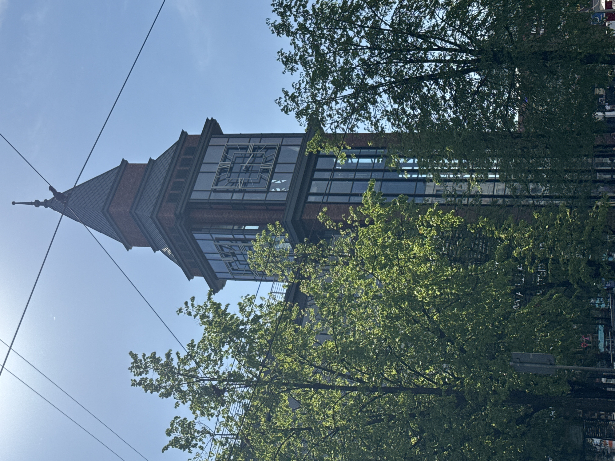 A low-angle shot of the clock tower of the Kuznechny Market in Saint Petersburg, Russia. The tower features a brick facade and large windowed sections. The view is partially obscured by a tree with bright green leaves. Power lines crisscross the upper left portion of the frame against a blue sky. A street light is visible to the right of the frame.