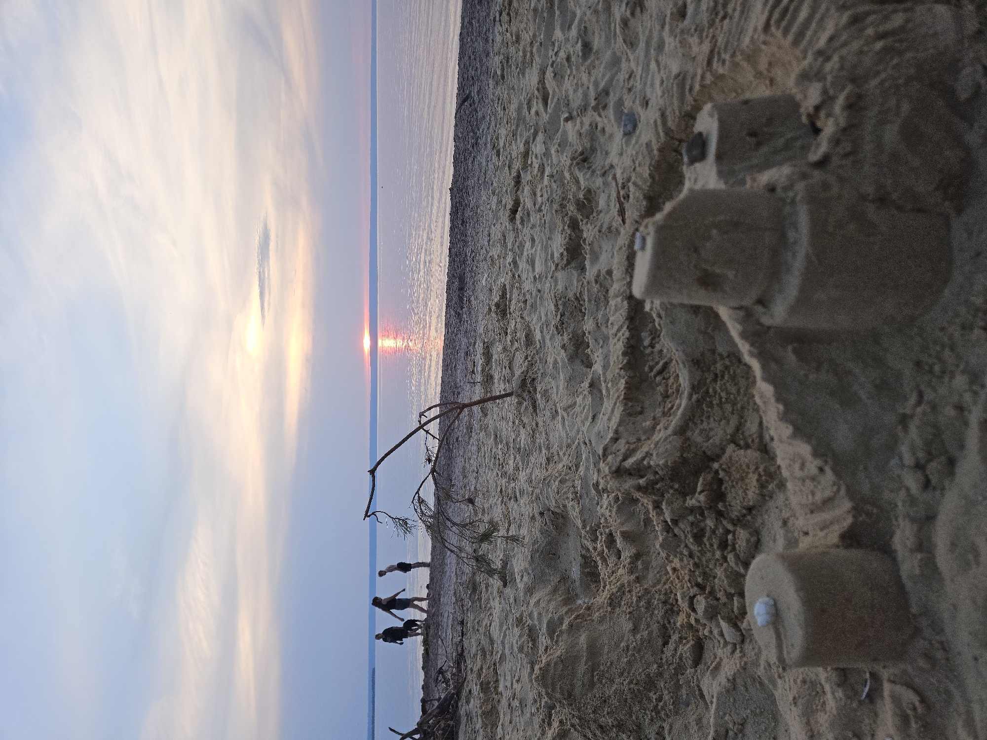 At sunset on a beach in Leninsky, Russia, three people and a dog walk near the water. The sky is a soft pastel blue and pink, with the sun a bright orb reflecting on the calm water. In the foreground, a sandcastle sits in the sand. A thin branch with a few needles from a pine tree is sticking out of the sand in front of the sandcastle. The beach appears to be a part of a large body of water, possibly a lake or sea, and there are some pieces of driftwood scattered on the sand. The overall atmosphere is serene and tranquil.