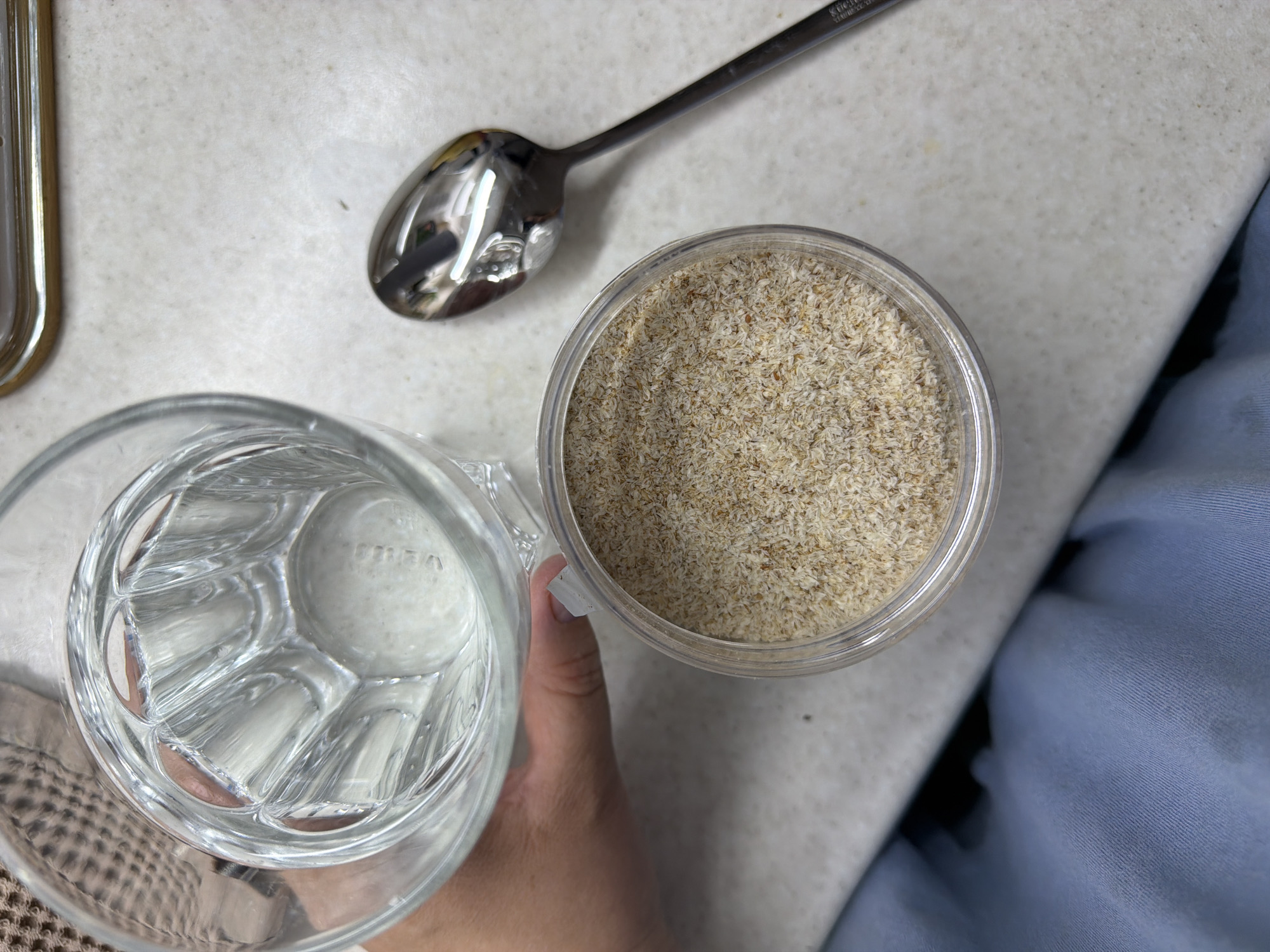 A top-down shot shows a glass mug filled with water held by a hand, a silver spoon, and a plastic container filled with psyllium husk. The scene is lit evenly and appears to be on a countertop. The location is not discernible from the image.