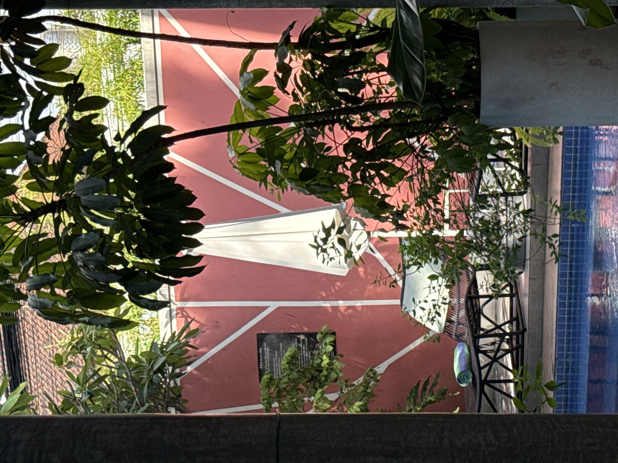 An eye-level, slightly rotated, outdoor photograph captures a scene near what appears to be a pool, featuring a red-colored patio area marked with white lines. Foliage from nearby trees and plants partially obscures the view. On the red patio, a white sunshade is partially visible, as is a dark informational sign posted on the patio surface. Adjacent to the patio is a blue-tiled wall or edge of a swimming pool. A dark metal chair with a rolled-up blue towel rests beside the pool. The image is likely taken in or around North Kuta, Indonesia, inferred from the metadata.
