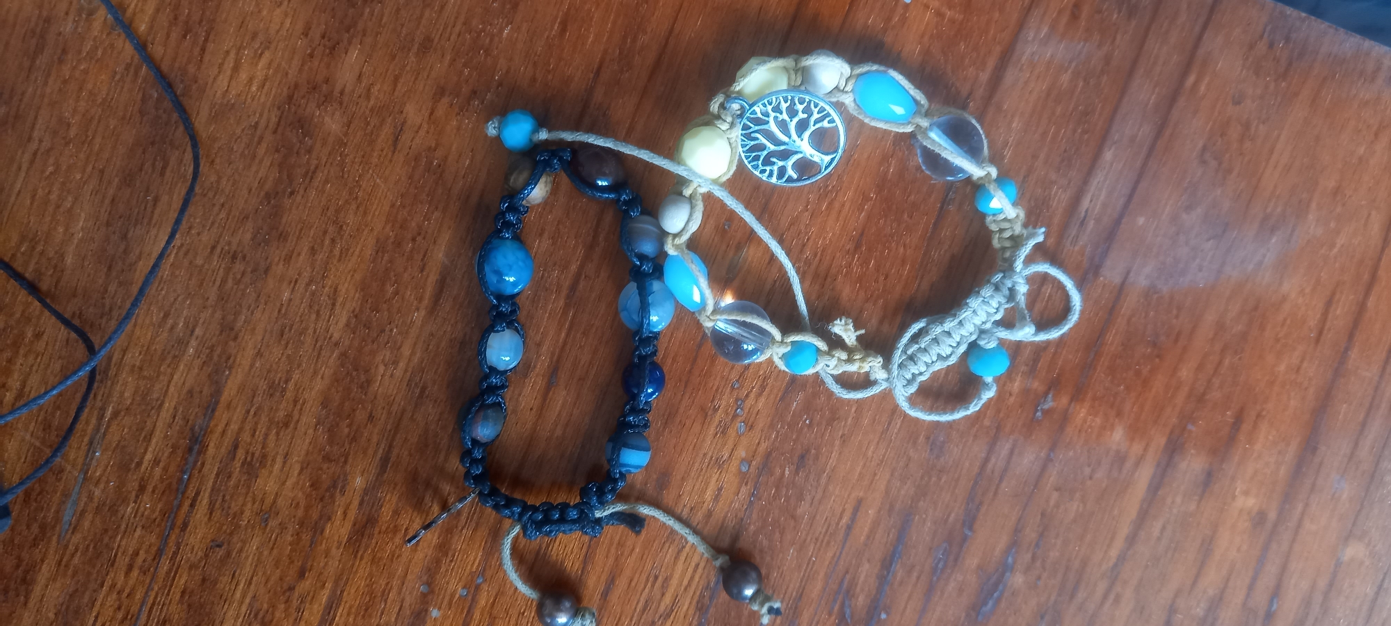 Two bracelets are displayed on a wooden surface. The top bracelet is black with blue and brown beads. The bottom bracelet is made of a lighter colored string and features clear, blue, and yellow beads, as well as a silver charm shaped like a tree. The wooden surface has a visible grain pattern and appears to be a table. The scene is likely indoors. There is no information to suggest this scene is located in Izhevsk, Russia.