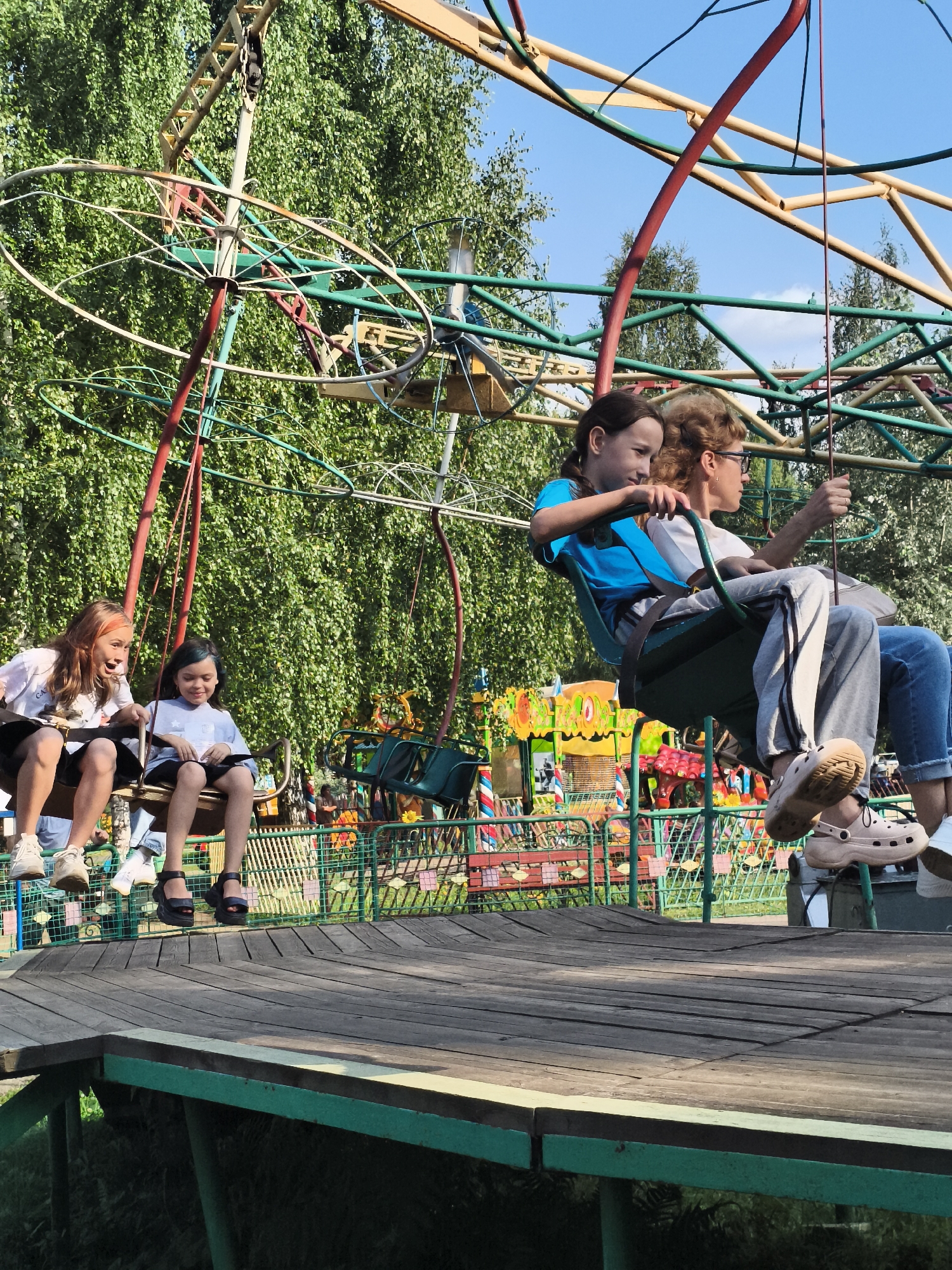 In Chaikovsky, Russia, a swing ride operates with several individuals enjoying the attraction. In the foreground, four people are seated in two swings, with two children on the left and two individuals on the right. The children on the left are a young girl with light brown hair, wearing a white t-shirt and black shorts, and a younger girl with dark hair, in a white shirt and dark shorts. On the right, a girl with dark brown hair in a blue t-shirt is seated next to a woman with curly blonde hair wearing glasses and a white t-shirt with gray pants. The ride itself is an elaborate metal structure with a wooden platform. In the background, a dense green forest provides a natural backdrop, with a bright blue sky overhead. The park appears to be operational, with other amusement park elements visible in the distance.