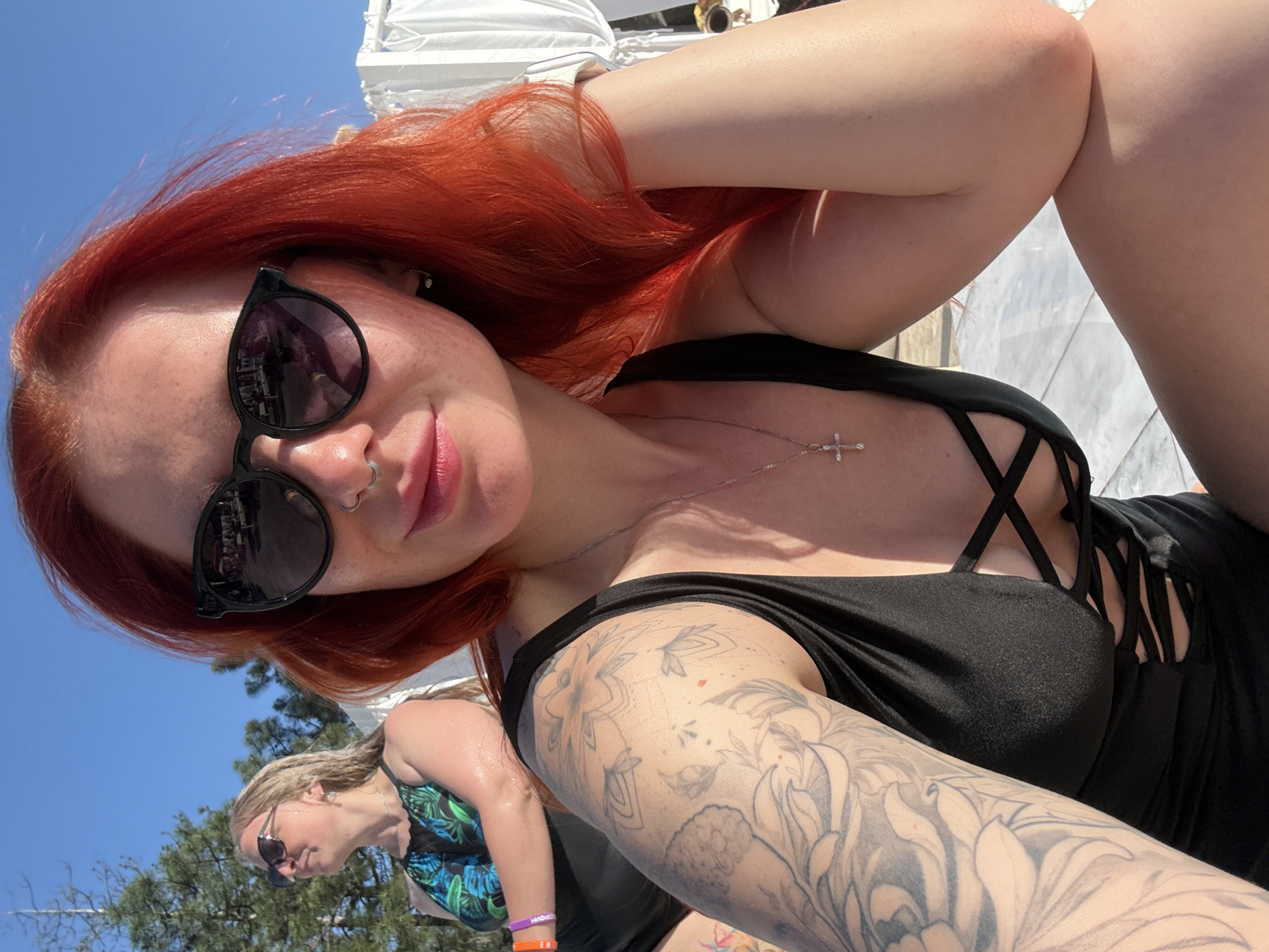 A woman with bright red hair is taking a selfie outdoors. She is wearing dark sunglasses and a black top with crisscross straps at the neckline. A silver necklace with a cross pendant is visible. Her left arm, which is tattooed with floral designs, is raised. A second woman is partially visible in the lower left corner of the frame. The setting appears to be a sunny outdoor location. Based on the image alone, it is not possible to determine if it was taken in Rostov-on-Don, Russia or to provide further forensic details.