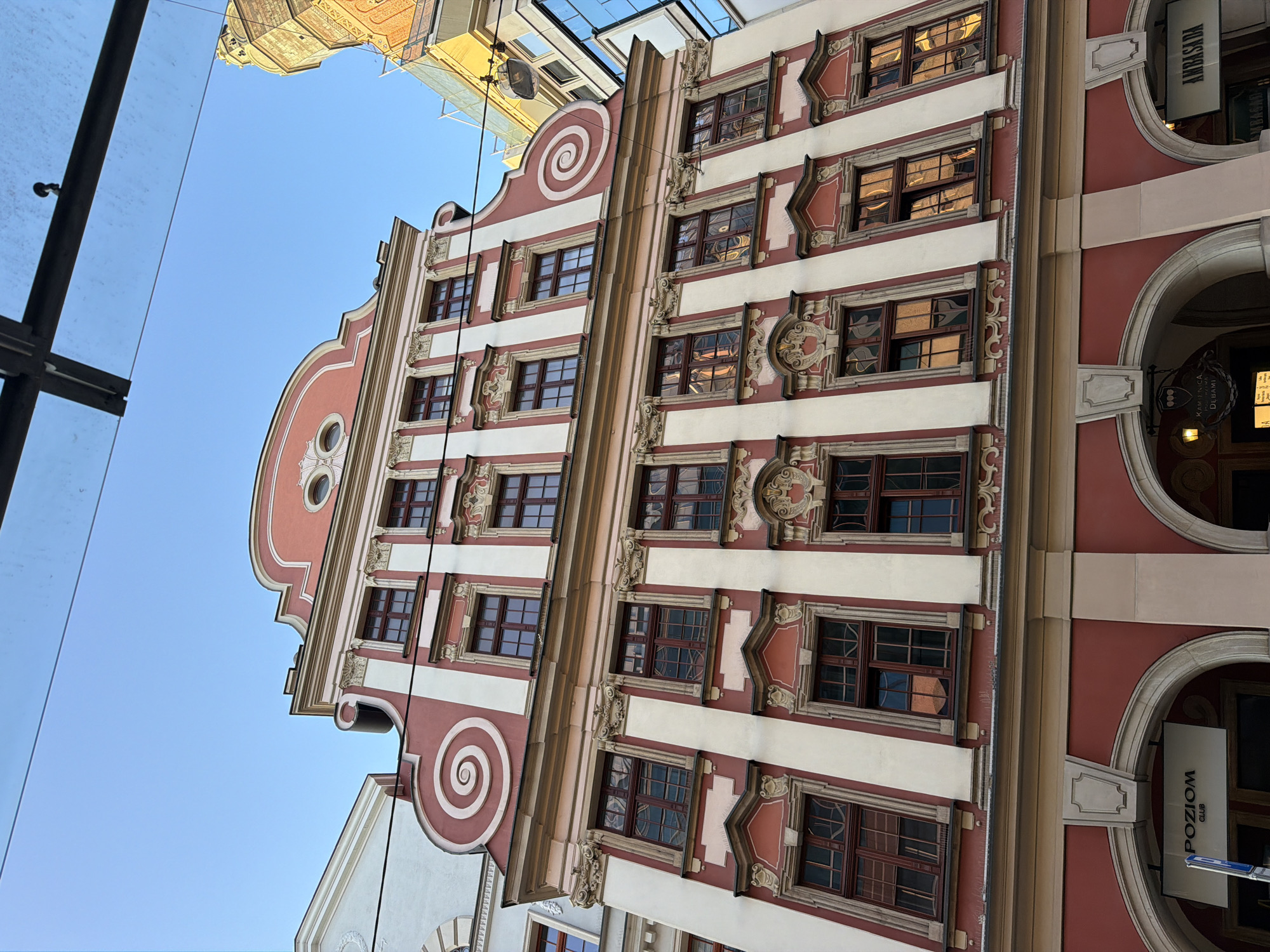 This is a low-angle, full-shot photograph of a row of historic buildings in Wrocław, Poland, likely taken on a clear day. The primary building, a pastel pink and cream facade with ornate detailing, dominates the frame. It features multiple stories with numerous windows, each framed by dark wood and featuring intricate mullions. The building's roofline is complex, with a curved pediment adorned with decorative scrolls and circular windows. To the left, a portion of a white building with a similar architectural style is visible, and to the right, a modern building with a glass facade and a brick tower under construction peeks into the frame. The street level of the primary building reveals arched entrances, with signage indicating "POZIOM CLUB" and "AMBASADA". Overhead, dark cables and a partial view of a glass and metal structure are visible. The sunlight casts reflections on the windows, hinting at the surrounding urban environment.