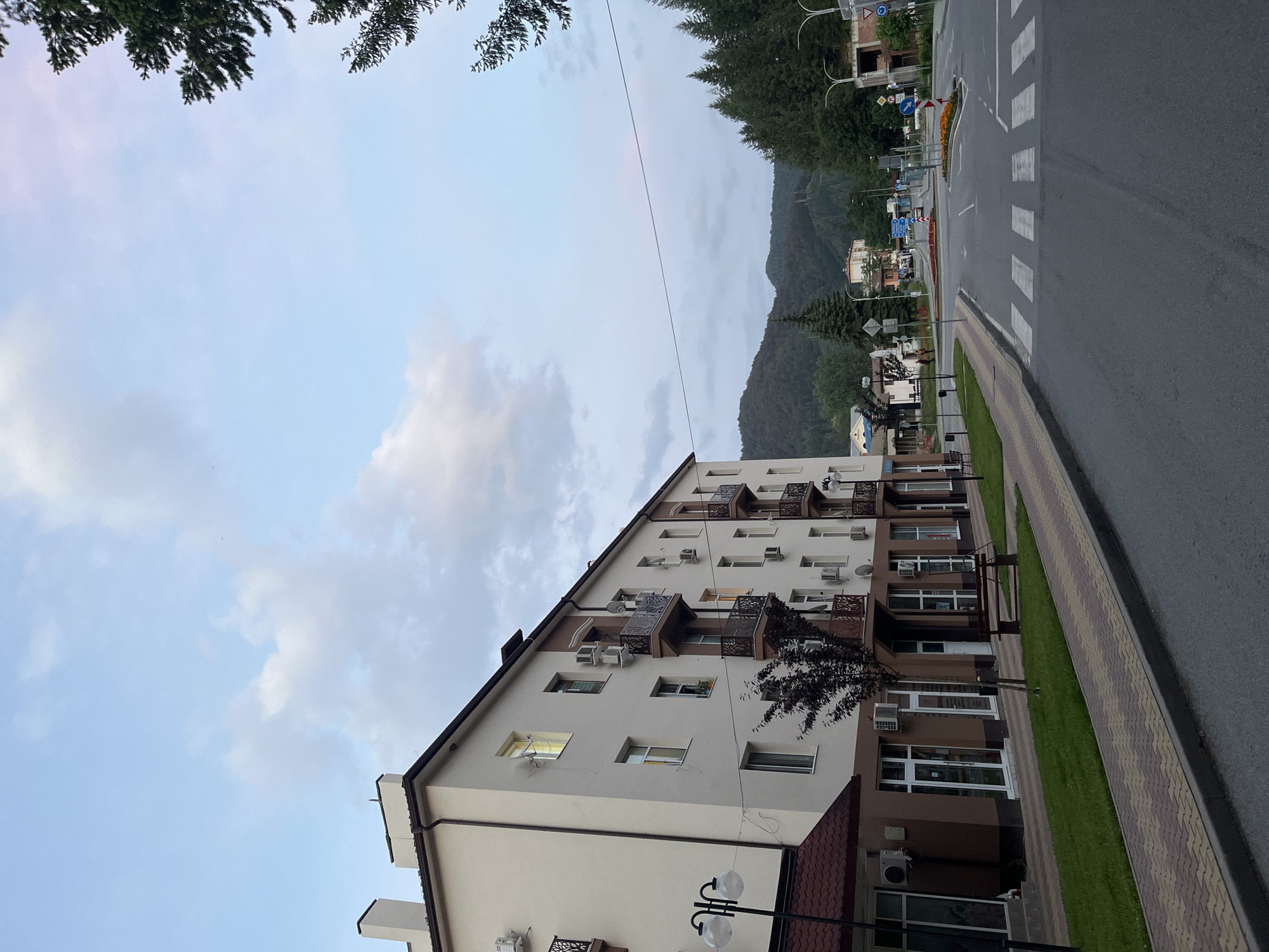 This image, oriented correctly, captures a street view in Rudozem, Bulgaria, with a blend of urban and natural elements. The main subject on the left is a multi-story apartment building featuring a modern design with white and brown facades, several balconies adorned with decorative metal railings, and numerous external air conditioning units.
In front of the building, a patterned sidewalk with a distinctive wavy design is visible, separated from the road by a narrow strip of green grass and a small tree. To the right, an asphalt road runs parallel to the building, featuring a clearly marked pedestrian crosswalk with white stripes. Along the far side of the road, several road signs are present, including a round blue sign indicating a mandatory right turn, though other signs are less discernible. Some construction appears to be underway in the distance on the right side of the road.
The background is dominated by dense, dark green coniferous forests covering rolling mountains, indicating a mountainous setting. The sky is a soft, light blue with scattered white and grey clouds, suggesting either an overcast day or early morning/late afternoon light. A thin power line traverses the sky horizontally across the middle of the frame. No people or explicit activities are visible, lending a calm and static atmosphere to the scene. The image showcases a residential area integrated into a natural, forested landscape.