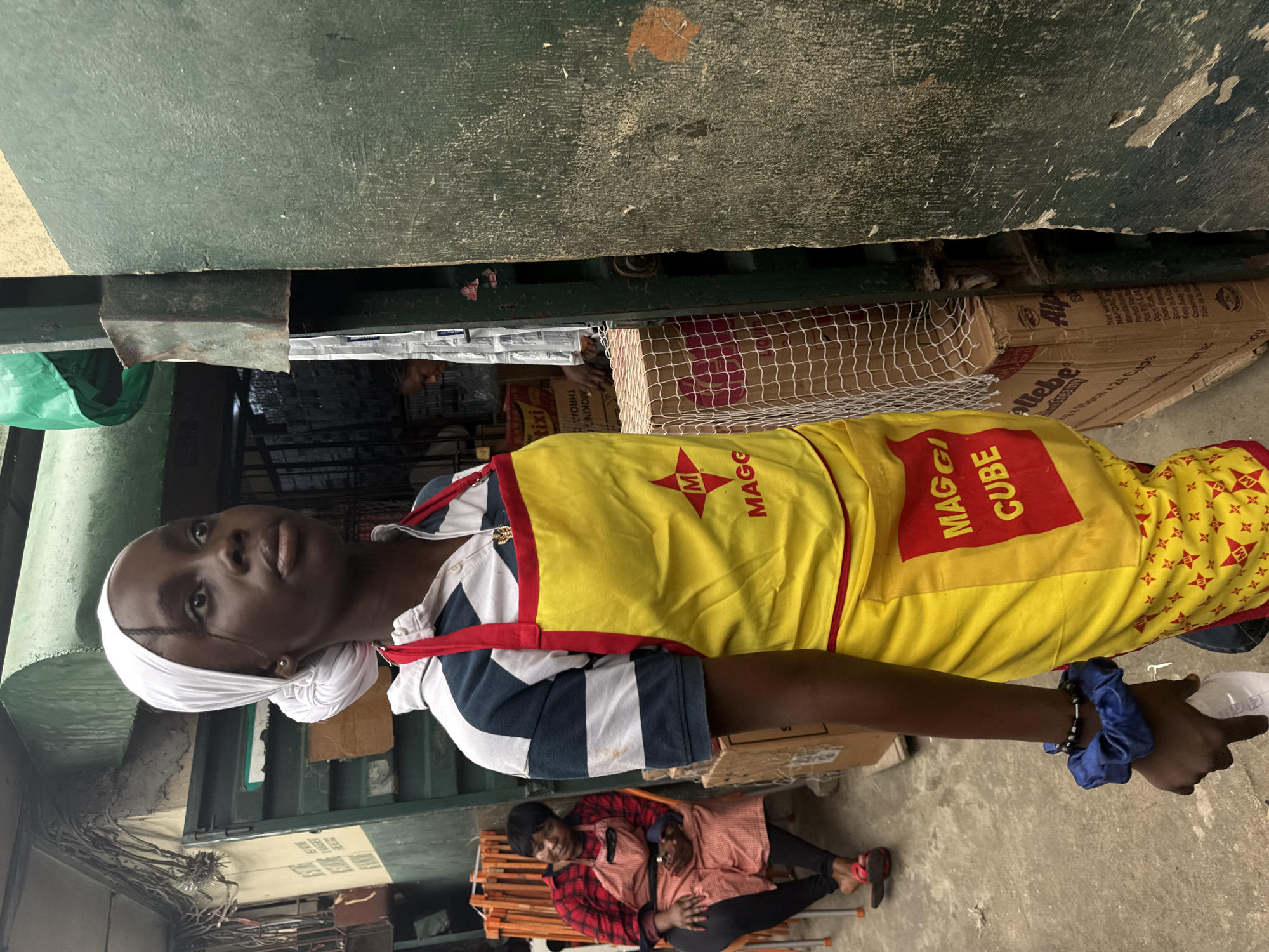 In a bustling marketplace in Lagos, Nigeria, a young woman stands near a stall, her gaze fixed upward. She wears a white headscarf and a striking yellow apron emblazoned with the Maggi Cube logo. Her striped shirt is partially visible beneath the apron. To her left, an older woman, also wearing an apron, sits on a chair, her arms crossed. Various boxes and merchandise are visible around them, hinting at the vibrant commerce of the location. The overall scene captures a snapshot of daily life in this busy urban environment.