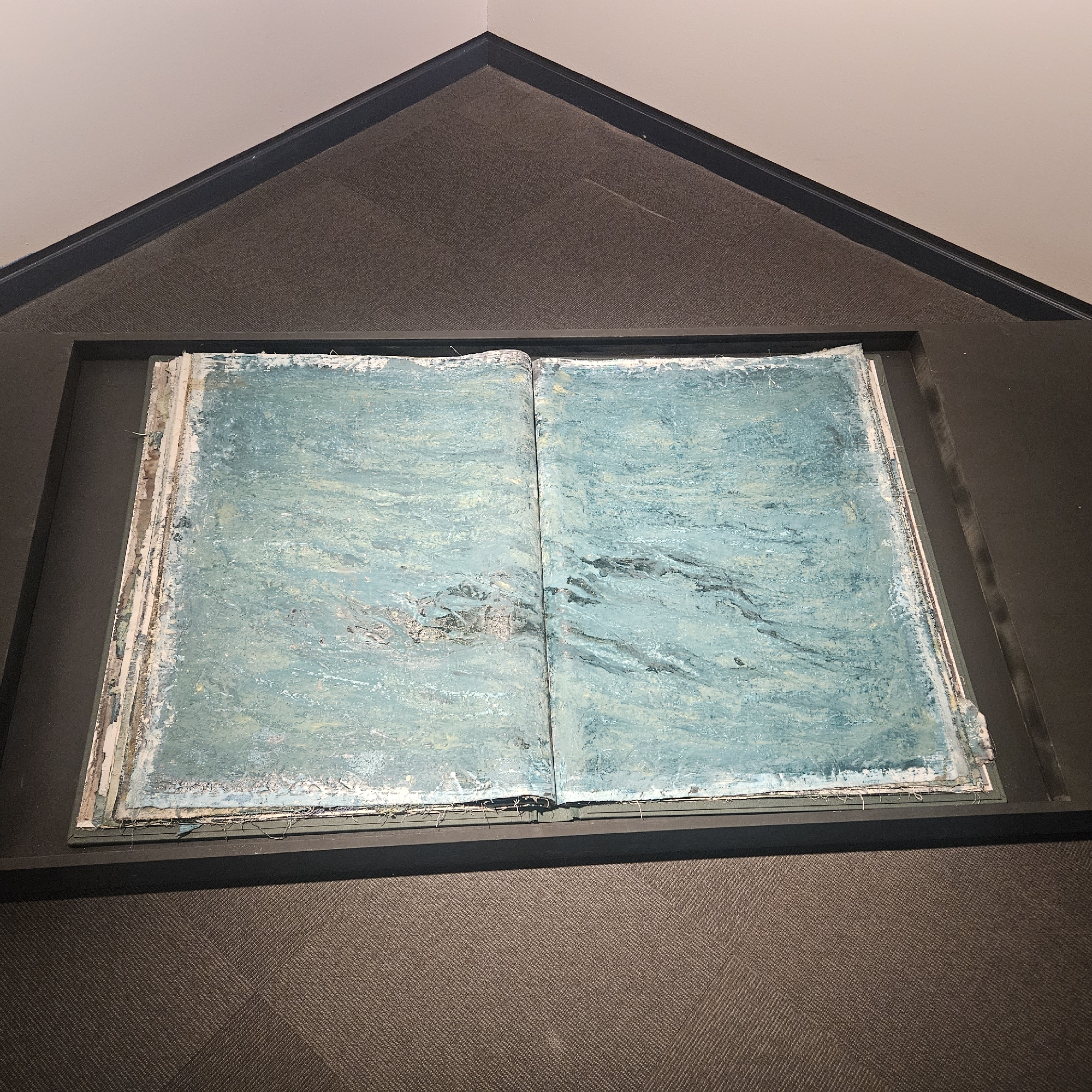The image shows an open book with heavily textured, abstract artwork on both pages. The artwork predominantly features shades of teal and blue, with visible brushstrokes, impasto techniques, and patches of white and darker, almost black, elements interspersed throughout. The pages themselves appear aged and somewhat distressed, with rough edges and visible layers of material or paper. The book is displayed open in a dark, recessed frame, which is mounted within a corner where two walls meet. The walls appear to be painted a light, neutral color, and the corner framing is black. Below the framed artwork, there is a dark, textured surface, possibly carpet or a similar material, on the floor. The lighting appears to be from above, casting subtle shadows. There are no people or animals present in the image. The location is identified as the city of Salt, Spain.