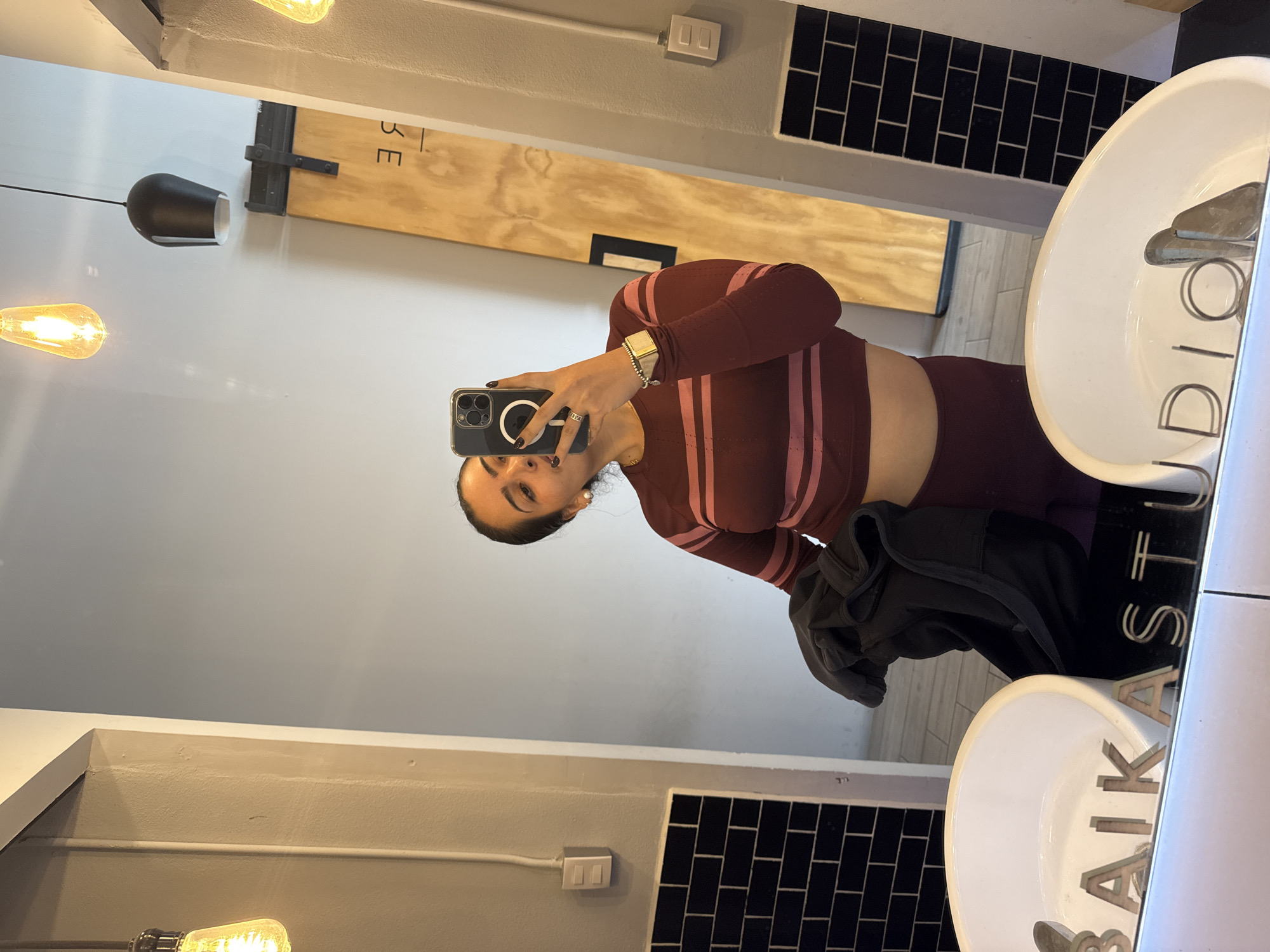 A woman is depicted taking a mirror selfie within what appears to be a studio changing room or restroom. She wears a long-sleeved, maroon athletic crop top with pink horizontal stripes, paired with matching maroon leggings. Her dark hair is pulled back, and she holds a dark jacket in her left arm. In her right hand, she holds a dark-cased smartphone with a visible MagSafe ring, positioned to capture the image. Her nails are painted dark, and a ring is visible on her right hand.
The reflection reveals two white oval sinks mounted on a light-colored counter. Below the counter, black subway tiles are installed. The name "BAIKASTUDIO" is visible in metallic relief letters on the reflective surface of the counter, between the sinks. The background walls are painted light grey. Overhead, two exposed Edison-style light bulbs and a black pendant light fixture provide illumination. To the right of the woman, a wooden barn-style sliding door is partially visible, displaying the reversed text "LIBRE." White electrical outlet or switch plates are present on the adjacent walls. This location is Baika Studio Libre in Puebla, Mexico.