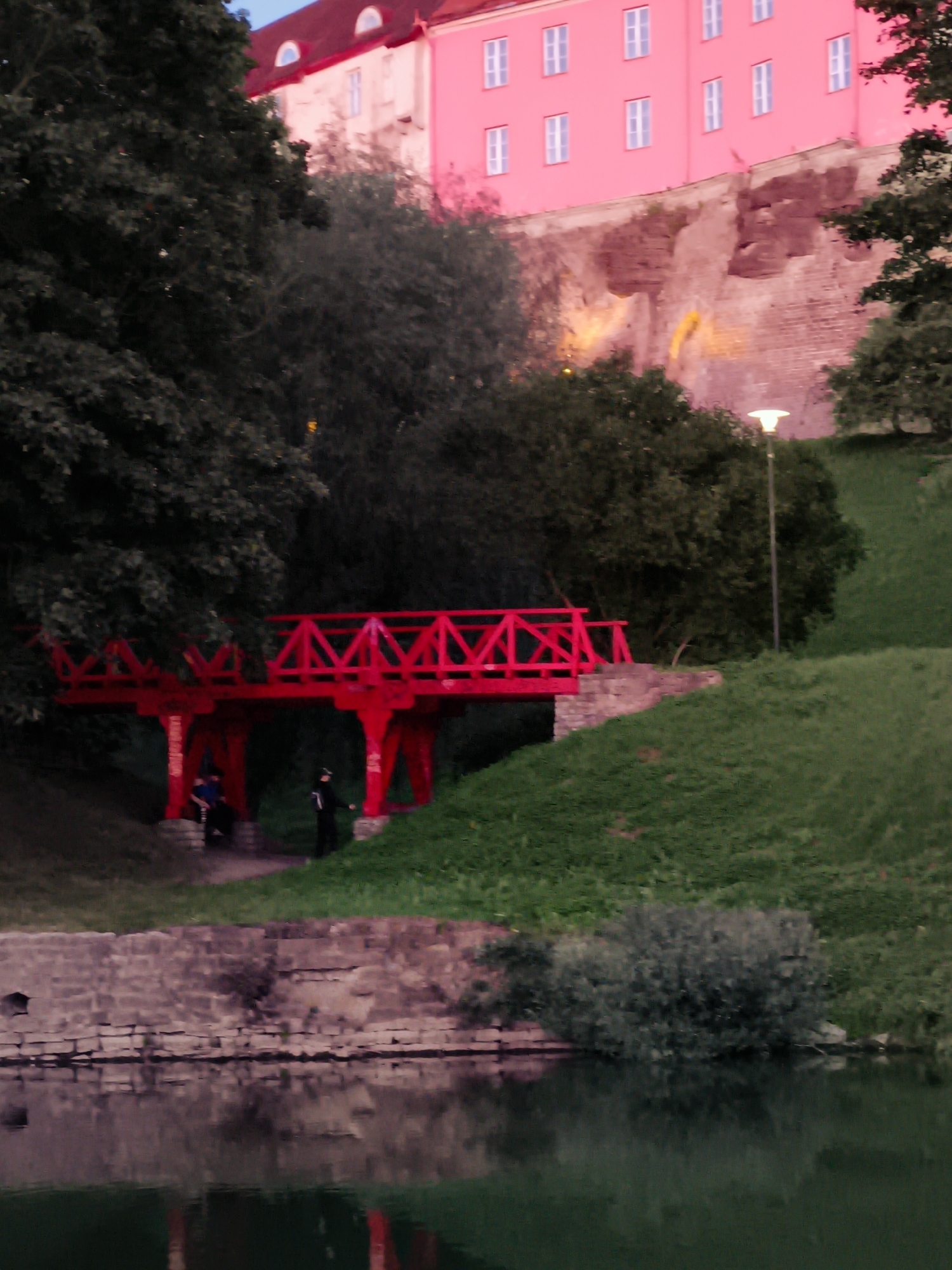 The image depicts a vibrant red footbridge spanning across a body of water in Tallinn, Estonia. The bridge, adorned with intricate geometric patterns, stands out against the lush green of a sloping park. In the background, a striking pink building with white-framed windows sits atop a stone embankment. Two individuals are visible near the bridge, one seated and another standing, adding a human element to the scene. The water reflects the surrounding environment, creating a serene and picturesque atmosphere. This bridge is located in Toompea Hill in Tallinn, known for its historical significance and scenic beauty.