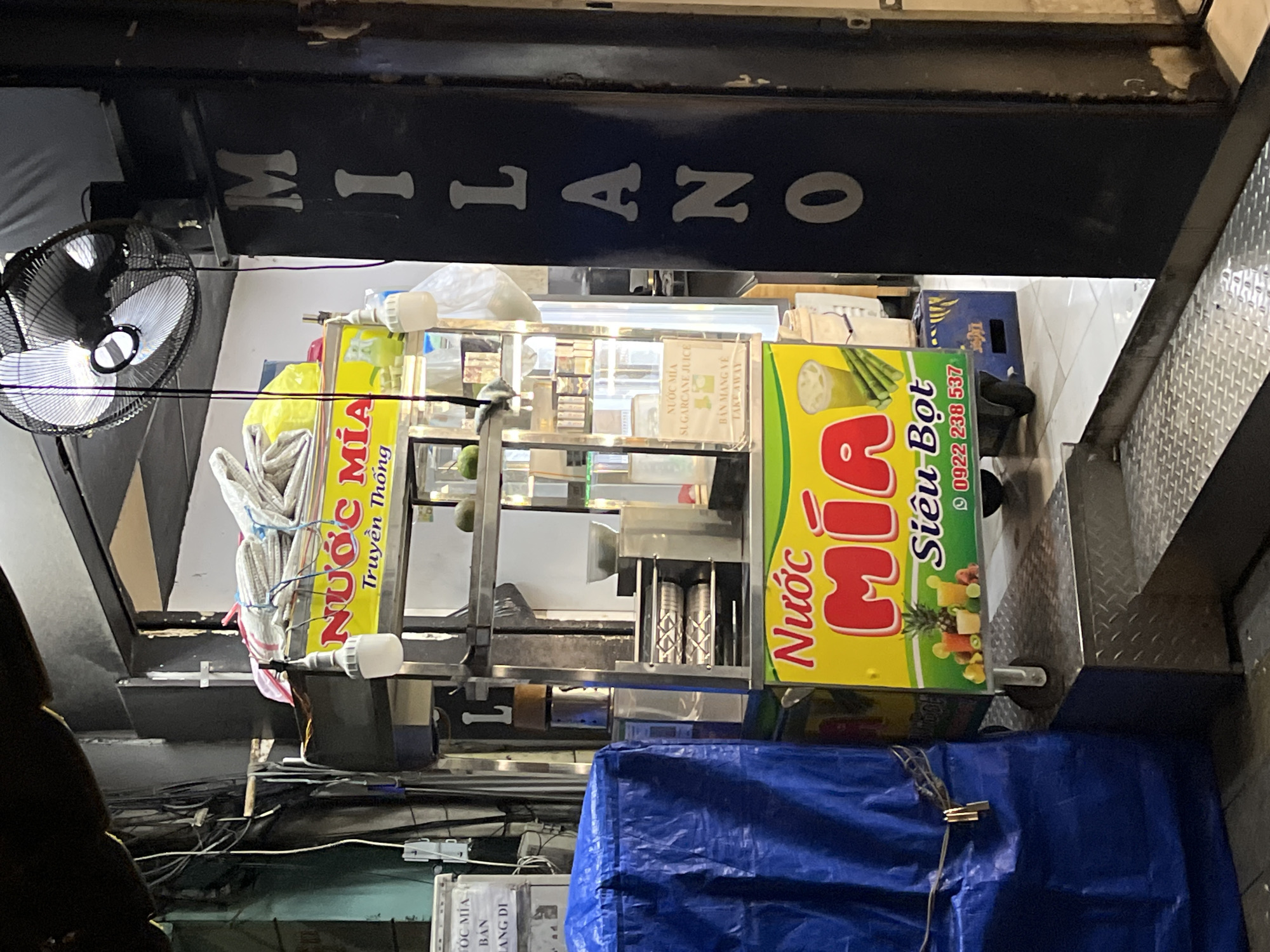 A street vendor cart in Thủ Đức, Vietnam, is positioned beneath a MILANO sign. A black fan with silver blades is mounted on the left. The cart has colorful yellow signage advertising NƯỚC MÍA sugarcane juice. A blue tarp covers the bottom of the cart. Visible cables and wires hang around the cart.