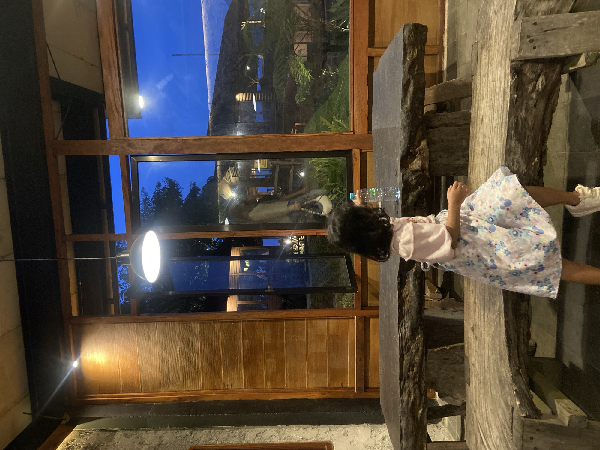 A young girl in a light-colored floral dress stands on what appears to be a rustic, wooden platform or seating area inside a building with large windows. She is near a table or countertop that has a water bottle on it. Through the windows, vegetation and what seems to be another building or structure can be seen. The location is Tamiajeng, Indonesia. The scene is illuminated by both interior lighting and natural light from the evening sky.