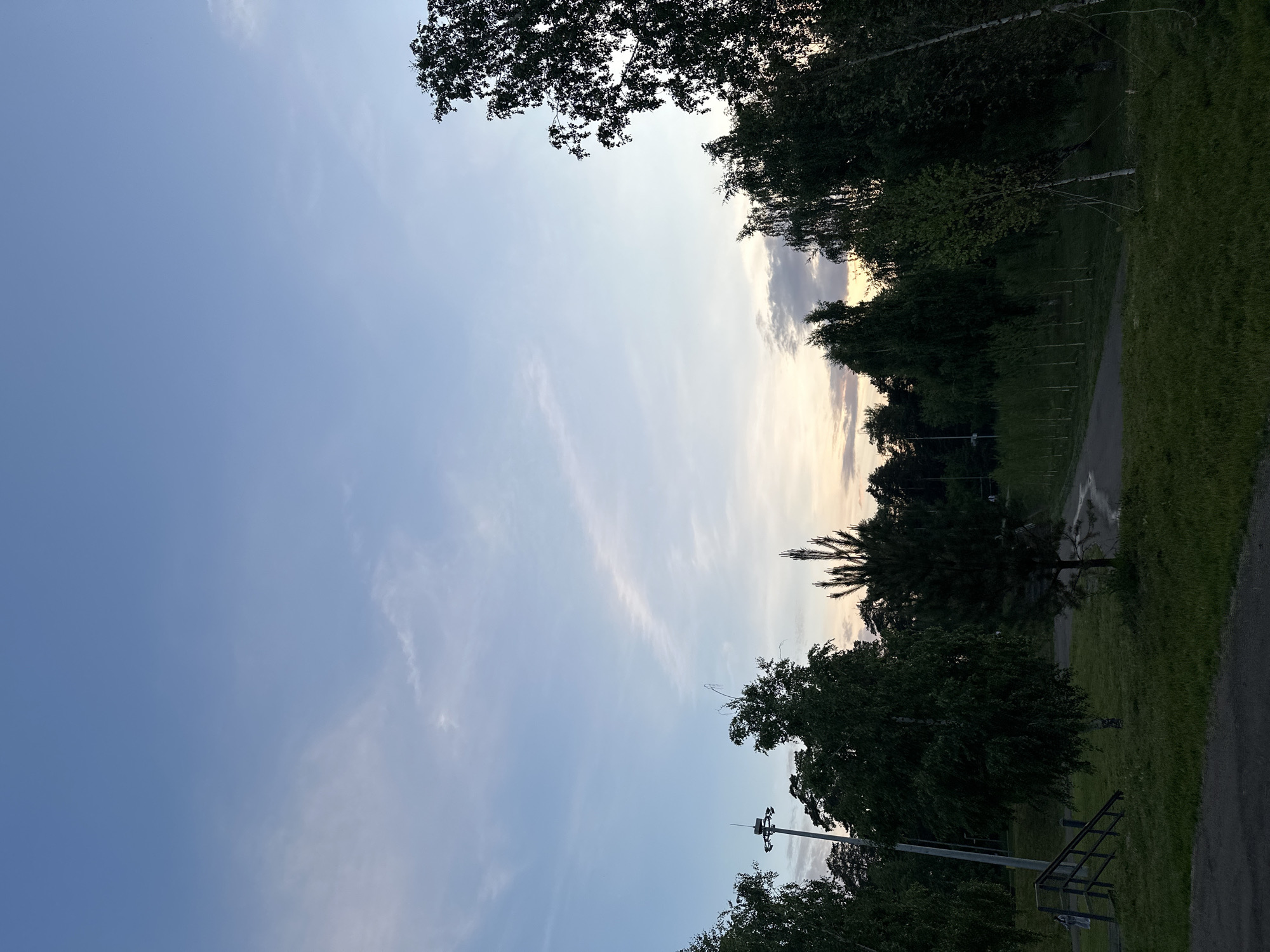 The image captures a twilight scene in a park-like area, likely in Kazan, Russia, given the provided context. The sky is a gradient of pale blue to a soft yellow and grey near the horizon, with thin, wispy clouds stretched across it. A solitary lamppost stands tall on the left side of the frame, its light extinguished. In the foreground, a dark asphalt path leads into a grassy area. Several trees, predominantly deciduous with dark green foliage, dominate the midground and frame the scene. A small, conical evergreen tree stands prominently in the center of the image, casting a shadow. In the distance, a dense line of smaller trees forms a verdant backdrop, with a few scattered buildings or structures barely visible through the foliage. A set of wooden stairs with a handrail is situated on the left, with two individuals walking away from the camera on a path below. The overall atmosphere is serene and natural, indicative of an outdoor recreational space during dusk.