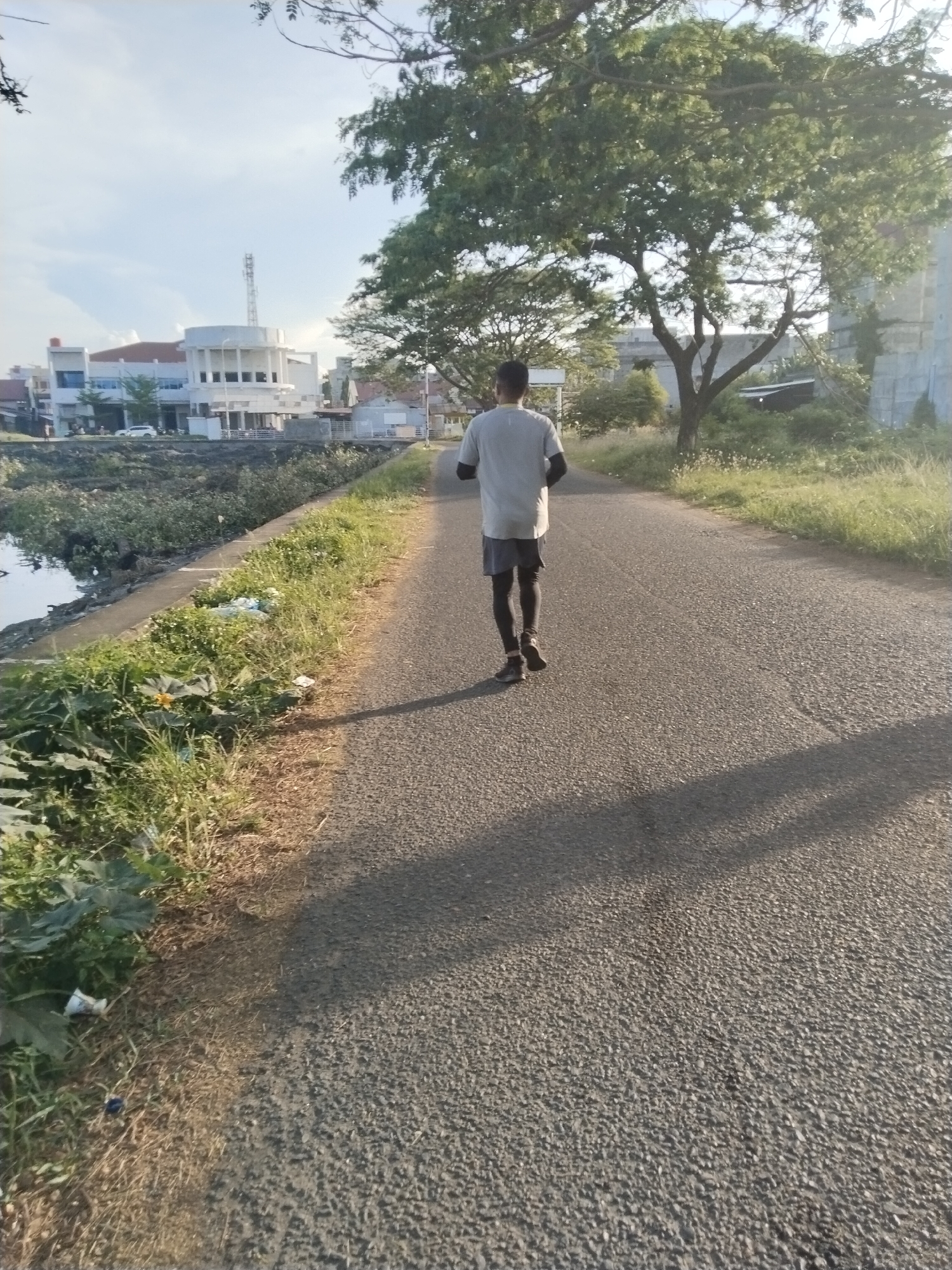 The image shows a man walking away from the camera on a paved road. He is wearing a light-colored t-shirt, dark shorts, and black leggings. The road is flanked by a waterway on one side and grassy vegetation on the other. In the background, there are buildings, including a white structure with a cylindrical section and a communications tower. A large tree provides shade on the right side of the road. The scene appears to be located in Lhokseumawe, Indonesia, based on the architecture and environmental features.