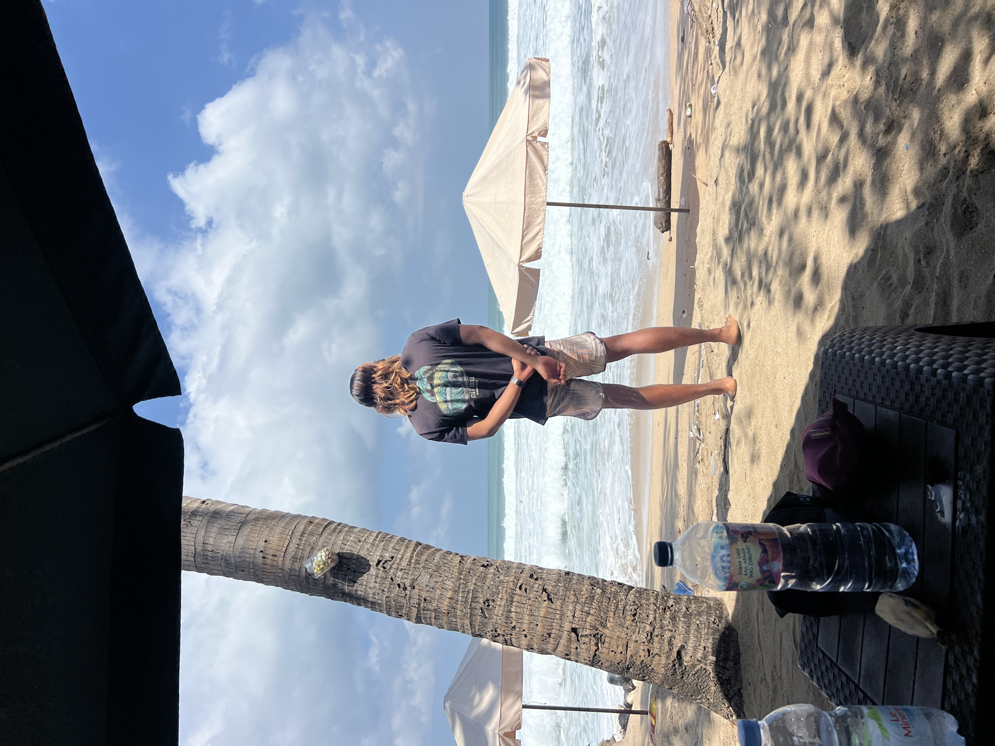 A person with long, wavy brown hair stands on a sandy beach in Kuta, Indonesia, facing the ocean. The person is wearing a dark t-shirt with a graphic design and patterned shorts. Their hands are clasped behind their back. In the foreground, a table holds two plastic water bottles and a dark-colored hat. To the left of the person, a large palm tree trunk stands prominently. Beyond the person, two beige beach umbrellas are set up on the sand, and the ocean waves are breaking onto the shore under a partly cloudy blue sky. A small plastic bag containing items, possibly snacks or trash, is attached to the palm tree.