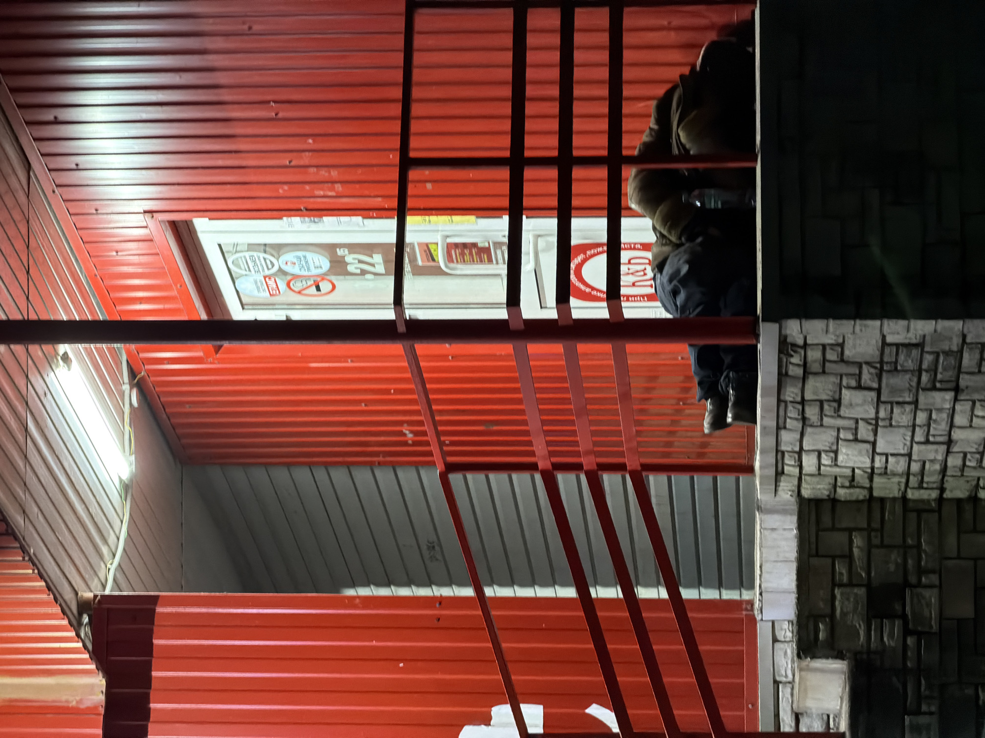 This nighttime image depicts the exterior of a commercial building in Chaikovsky, Russia. The structure features bright red corrugated metal siding. An individual is lying prone on a raised stone platform situated to the right of a white glass entrance door. The person is covered in dark clothing and a brown blanket or similar material, with dark shoes visible at their feet. A clear plastic bottle is present on the platform near their upper body.
A dark red metal railing extends across the foreground, partially obscuring the individual. The white glass door displays multiple circular decals, including a "No Smoking" symbol and the numerical "22¹⁵," likely indicating operating hours. Russian text, specifically "К&Б" (Krasnoe & Beloe), is also visible on the door's glass. Overhead fluorescent lighting illuminates the area directly above the entrance. The base of the building is constructed from patterned grey and white stone blocks. To the left of the entrance door, behind the railing, a section of grey corrugated metal siding is visible.