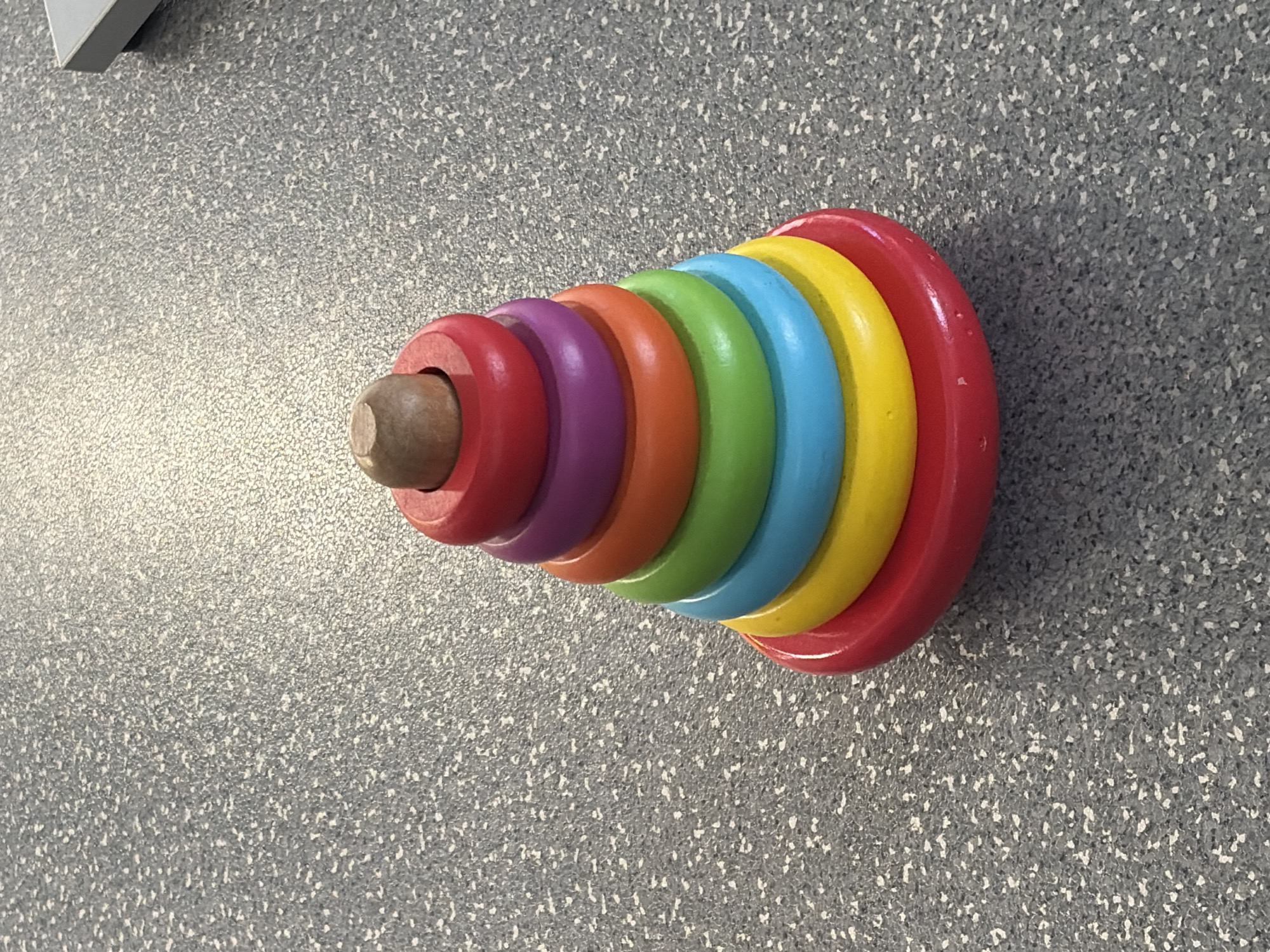 A wooden stacking toy, featuring rings of red, purple, orange, green, blue, and yellow stacked on a red base, with a small wooden peg at the top, is positioned on a speckled gray surface. The toy is the sole focus of the image, with no people or other objects clearly visible in the frame. The lighting suggests an indoor setting, possibly a home or educational environment. There is no information available in the image to determine its location in Teplice, Czechia.
