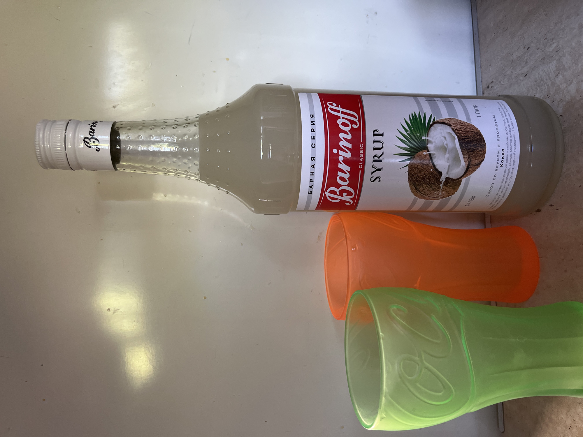 A bottle of Barinoff Coconut Syrup stands upright, positioned on a flat surface next to two plastic cups. One cup is lime green and appears to be a Coca-Cola branded glass, while the other is a solid orange color. The background is a plain, off-white, vertical surface, possibly a wall or cabinet. The scene is lit from the front, casting subtle shadows. The image does not contain any indications of being in Shymkent, Kazakhstan, or any other specific geographic location.