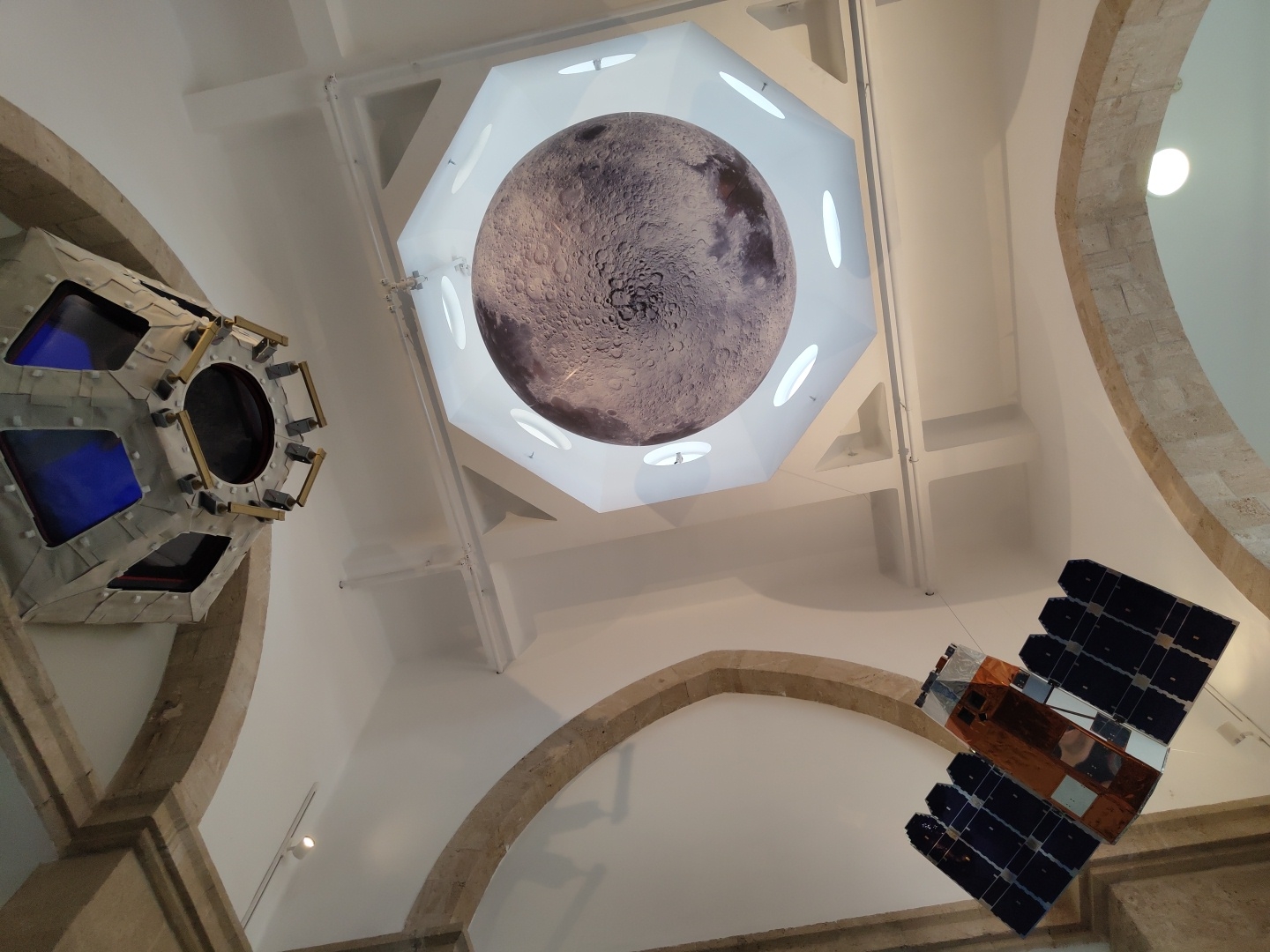 The image captures an indoor scene, likely a museum or exhibition space, with a focus on astronomical models. In the center of the frame, a large, detailed model of the Moon is suspended from the ceiling. The Moon model is illuminated from above by an octagonal structure with recessed lighting. To the left, a metallic, multi-faceted object resembling a spacecraft or satellite module is suspended. It features several windows with blue interiors and circular openings. In the lower right, another model, clearly a satellite with solar panels, is hanging. The architecture suggests an older building, with curved, stone arches visible in the foreground and on the right side of the image. The walls and ceiling are painted white. The overall impression is that of an exhibit dedicated to space exploration. This scene is located at the Madatech, The Israel National Museum of Science, Technology, and Space, in Haifa, Israel.