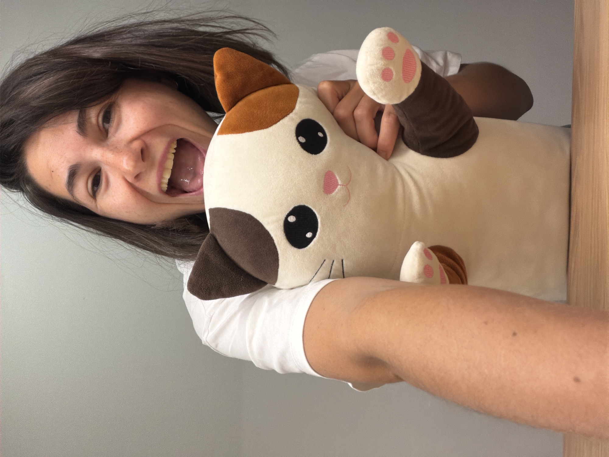 A woman with dark hair and a wide smile is holding a plush cat toy. She is wearing a white t-shirt. The cat plush toy is cream-colored with brown and orange patches. It has large, black, round eyes and a pink nose. The background is a light gray wall with a wooden doorframe on the right. The location (Budva, Montenegro) is not evident in the image.