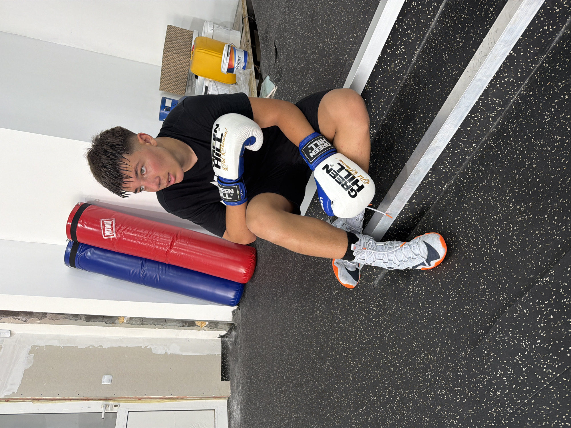 A young male boxer, wearing a black t-shirt, black shorts, grey and orange boxing shoes, and white and blue boxing gloves, is sitting on a metal platform in what appears to be a boxing gym. Two large, deflated punching bags, one blue and one red, are leaning against a wall in the background. There are also several paint cans and containers on a wooden pallet. The floor is black with white speckles, typical of a gym mat. The overall scene suggests a training or preparation area for boxing, possibly in a location in Blagoevgrad, Bulgaria.