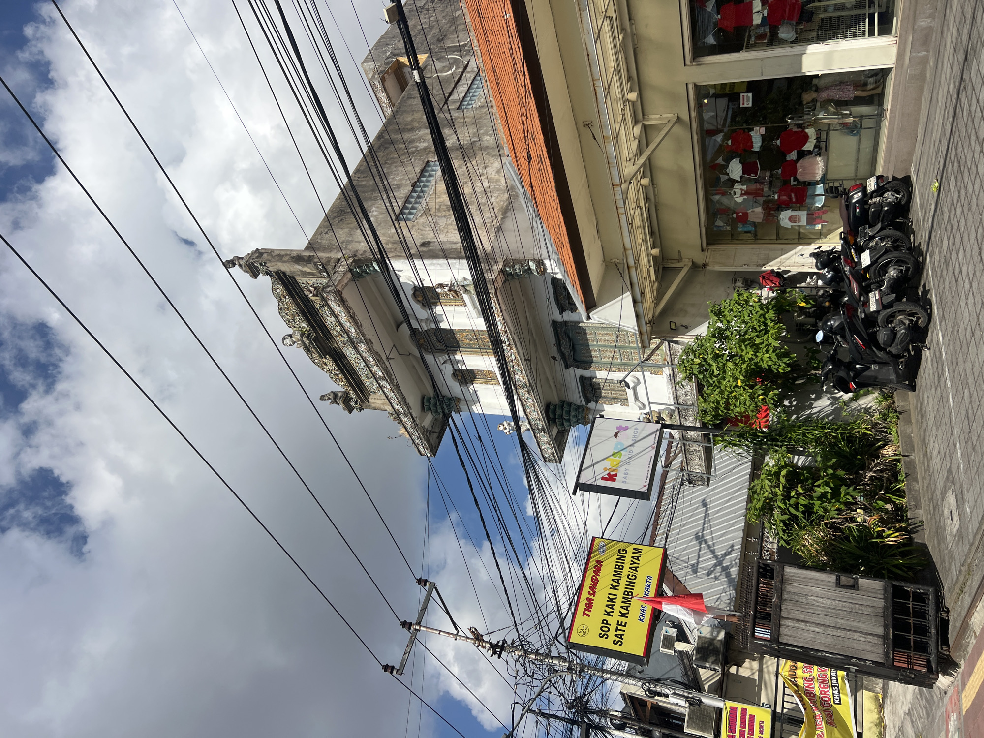 The image depicts a street scene in Denpasar, Indonesia, under a cloudy sky. In the foreground, several parked motorcycles are visible on a paved surface. Adjacent to the motorcycles is a shop with a glass storefront displaying children's clothing and a mannequin. Above the shop, a building with an ornate facade and tiled roof is partially visible. Further to the left, a yellow sign advertising "Tiga Saudara" (Three Siblings) restaurant with Indonesian text is mounted on a pole, along with a complex network of electrical wires. A red and white Indonesian flag flies near the restaurant sign. Another sign above the entrance to the restaurant reads "Kids Shop". The buildings appear to be a mix of traditional and modern architecture, characteristic of urban areas in Bali.