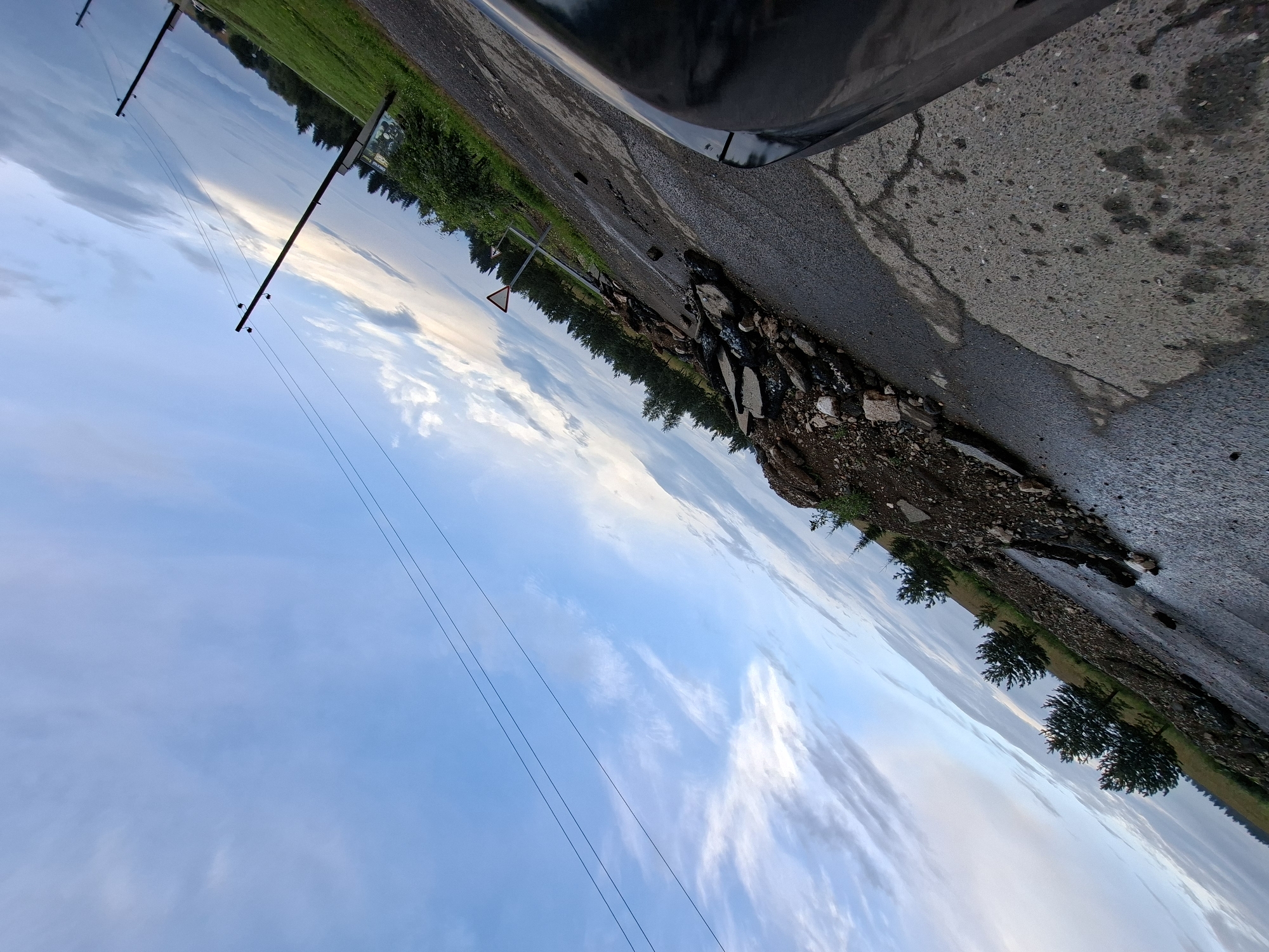 The image depicts a road partially obstructed by debris, possibly from a landslide. The road surface is cracked and wet in places. Several trees are visible along the side of the road, and a grassy field extends beyond them. The sky is blue with scattered clouds, and power lines are visible crisscrossing the upper portion of the frame. There are also two traffic signs on the right side of the road. The image appears to have been taken from a vehicle, as the hood of a car is visible in the foreground. The scene is outdoors, under a twilight sky. There is no indication of the city of Yugo-Kamskiy, Russia in the image.