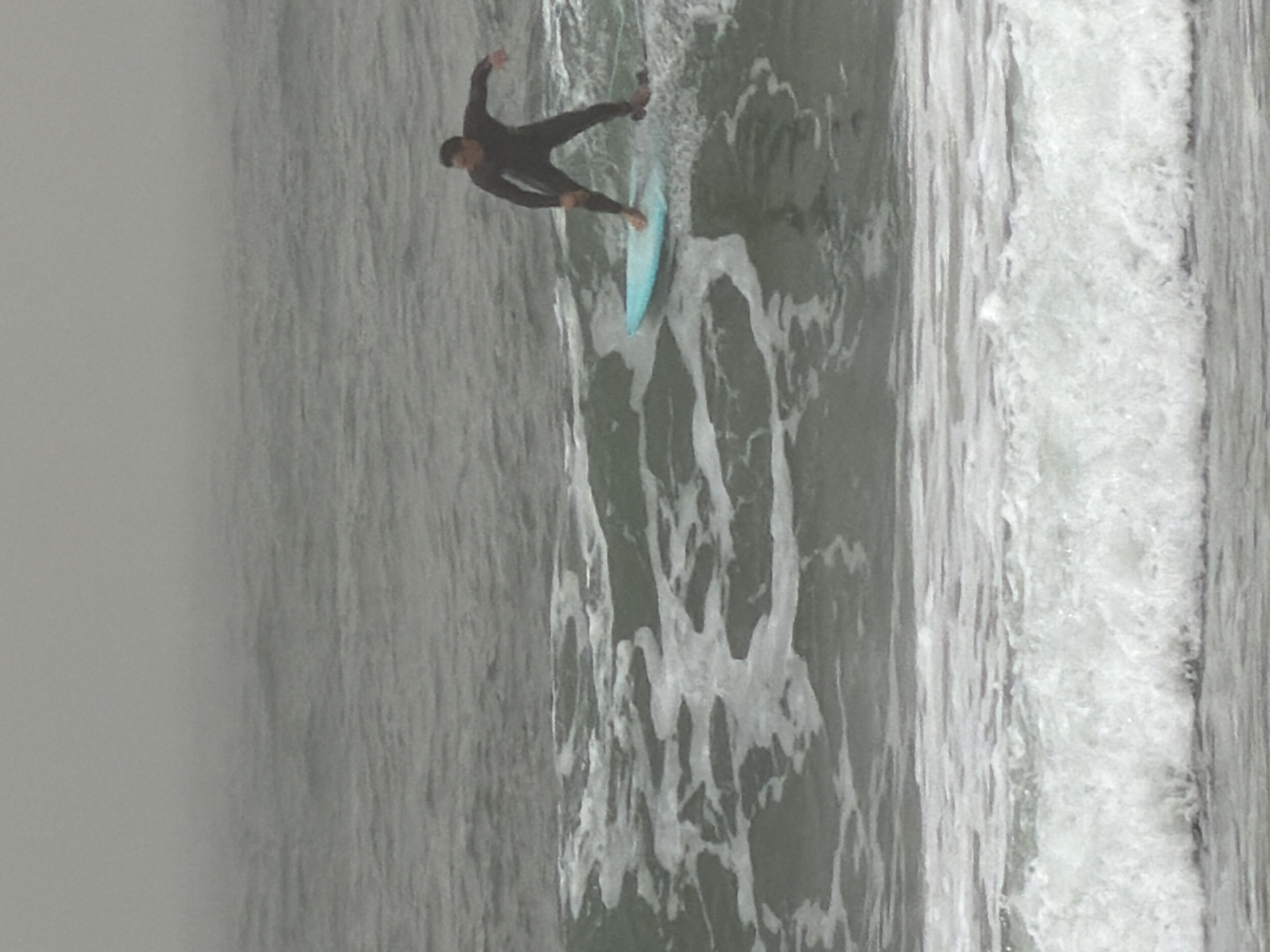 A surfer in a dark wetsuit is riding a light blue surfboard on a wave. The sea is choppy with white foam. The background is foggy and grey. This scene may be located in Tel-Aviv, Israel.