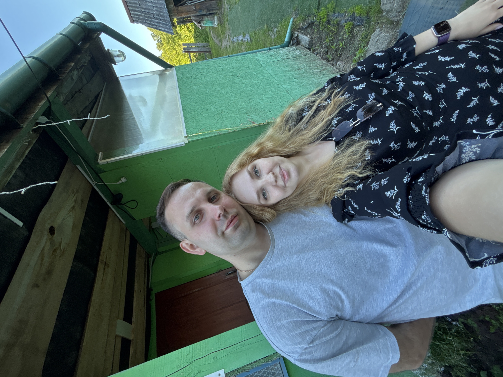 A man and a woman are captured in a selfie in Rozhdestveno, Russia, standing in front of a rustic green structure. The man, positioned on the left, has short dark hair and light eyes, wearing a light gray short-sleeved t-shirt. The woman, on the right, has long wavy blonde hair and light eyes, wearing a black dress with white floral patterns, sunglasses tucked into her neckline, and a purple smartwatch on her left wrist. Her head rests on the man's right shoulder as both look towards the camera with gentle smiles.
The background features a bright green wooden building or annex. Its exterior walls are constructed from both horizontal wooden planks and textured paneling, possibly OSB. A dark brown wooden door is visible on the left side of the structure, and a small white electrical outlet is mounted on the green wall near the man's left ear. On the right side, a clear corrugated plastic sheet serves as a window or light panel. Above, a wooden overhang reveals exposed beams and black sheeting. A dark green metal gutter system runs along the edge of the roof, with a corresponding green downspout extending vertically down the right side of the building. A globe-shaped light fixture is affixed to the underside of the overhang, and a string of white, unlit decorative lights hangs down.
Beyond the green structure, a narrow dirt path winds into the distance, flanked by patches of green grass and scattered rocks. Further back on the right, a smaller, older wooden building with a gray corrugated roof is partially visible. The scene is set outdoors under a clear blue sky with ample natural light, and green foliage, including trees and bushes, fills the horizon.