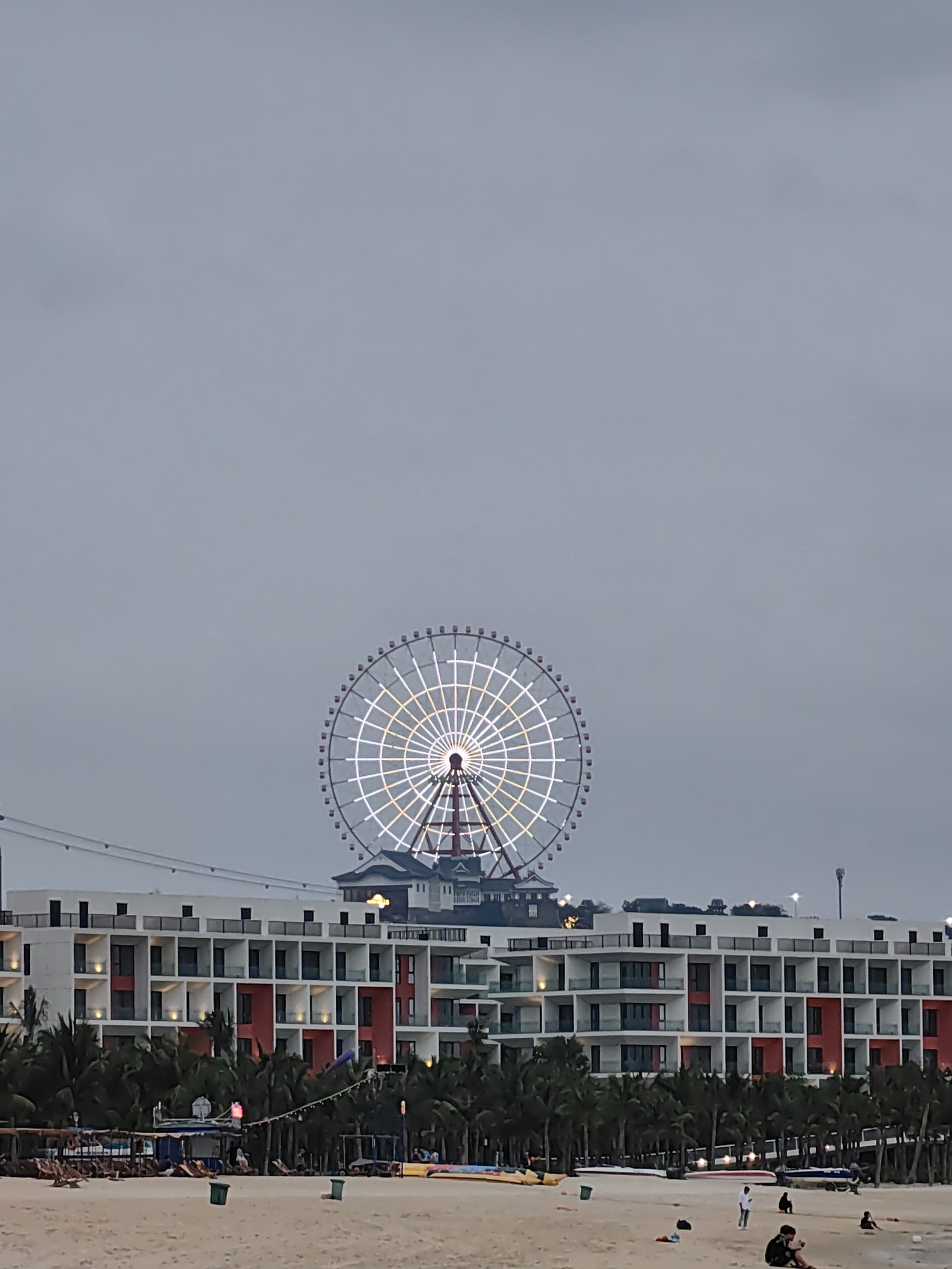 This image captures an evening scene at a coastal resort, likely in Bai Chay Ward, Vietnam. A large, brightly illuminated Ferris wheel dominates the background, its intricate white and golden lights glowing against a dark, overcast sky, suggesting dusk or night. Below the Ferris wheel, a substantial, modern multi-story building stretches across the mid-ground. This structure, possibly a hotel or resort, features a contemporary design with white facades, numerous balconies, and striking red-colored sections.
In front of the building, a line of palm trees partially obscures its lower levels. The foreground consists of a wide, sandy beach where several individuals and small groups are visible. These people appear to be relaxing, with some seated on the sand and others standing. Various beach items, such as what resemble small boats or kayaks and lounge chairs, are scattered along the shoreline. Two green trash cans are also visible on the sand. Faint lines, possibly power cables or amusement park infrastructure, traverse the upper portion of the building. The overall impression is one of leisure and evening entertainment at a popular tourist destination. No discernible text is visible in the image.