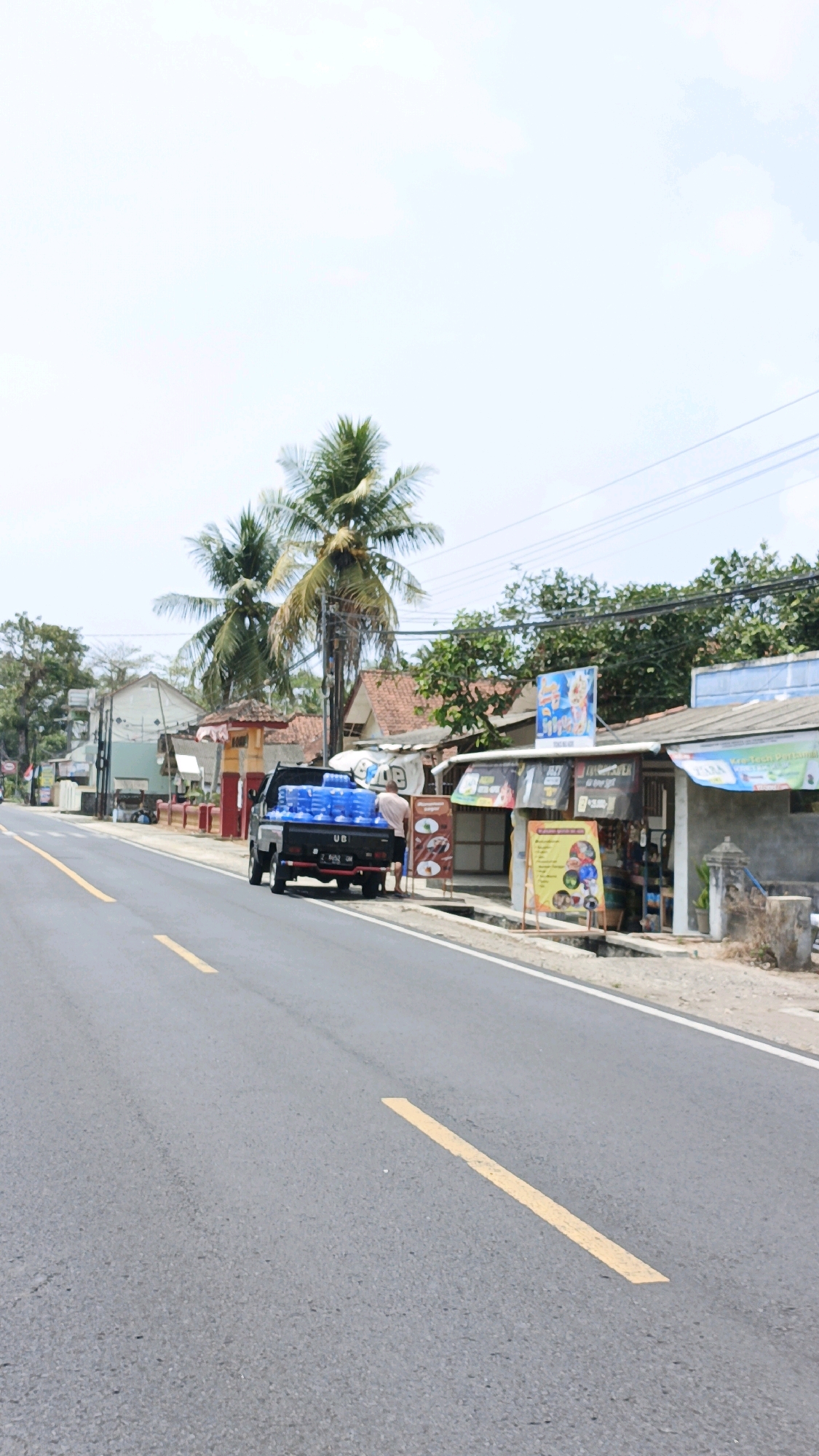 The image depicts a roadside scene in Sukaresik, Indonesia, featuring a paved road with yellow lane markings. A black pickup truck is parked on the right side of the road, laden with blue plastic water bottles. A person, partially visible, stands near the rear of the truck. Adjacent to the road is a row of small buildings or stalls, some with signs and awnings, suggesting a commercial area. Tropical vegetation, including palm trees, is visible in the background, with a bright, overcast sky above. The environment appears to be a rural or semi-urban setting.