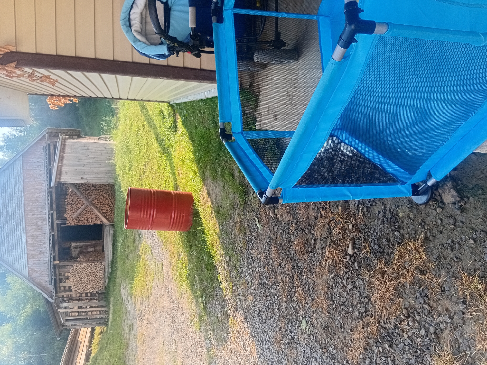 This is a medium-angle, outdoor shot taken in Verkhniachka, Ukraine. The foreground features a blue mesh playpen. To the right of the playpen, a stroller with a blue canopy is parked. Behind the playpen and stroller, a red barrel stands on a grassy area. In the background, a wooden structure, possibly a shed or barn, is visible, with stacked firewood in front of it. The ground is a mixture of gravel and grass. Trees and foliage are present in the far background. The lighting suggests it is daytime.