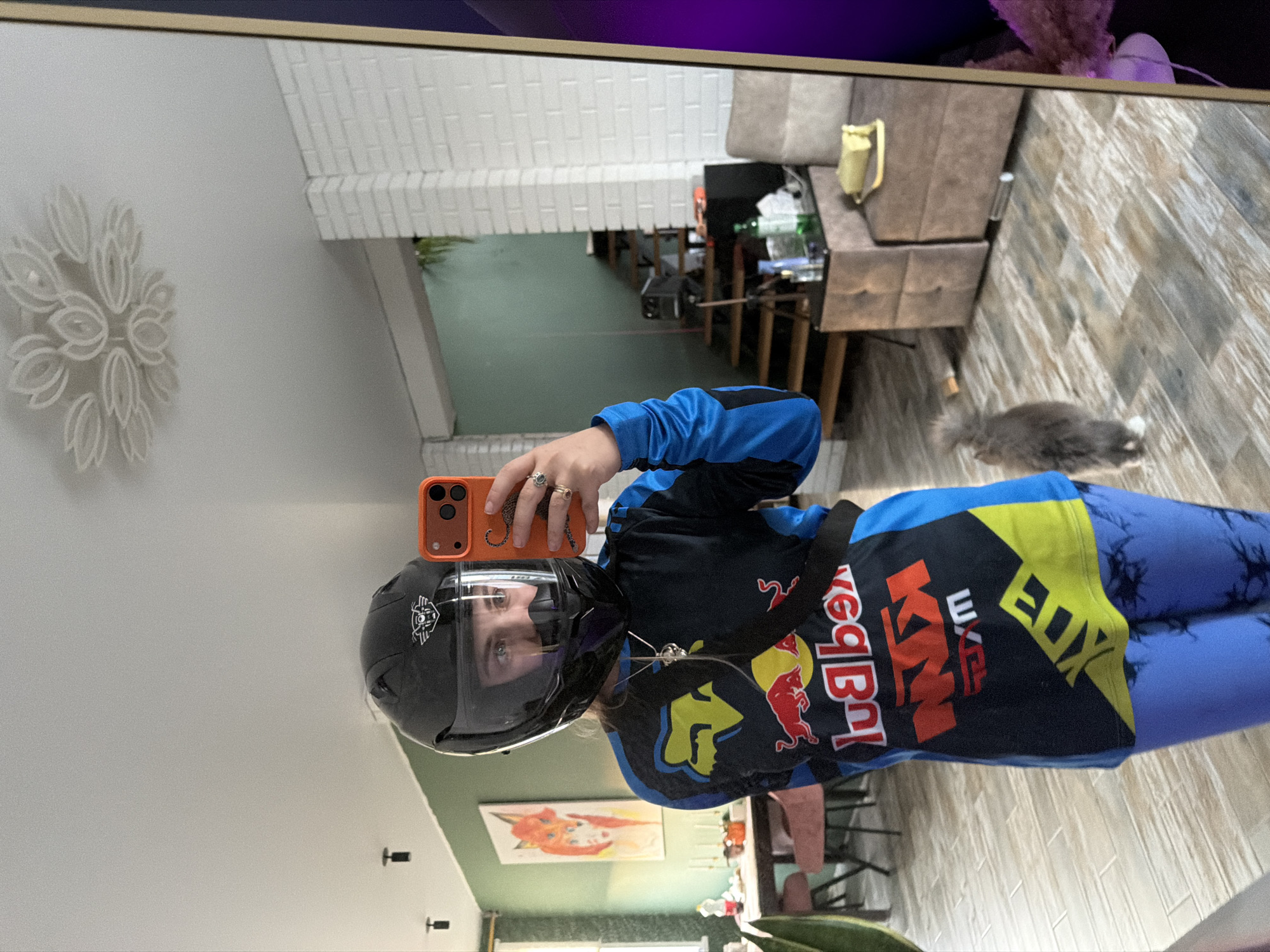 The image appears to be a mirror selfie taken indoors. A person wearing a motorcycle helmet, a racing shirt with "Red Bull KTM" logos, and blue leggings is holding an orange smartphone. The reflection shows a room with a staircase in the background, a gray cat near the bottom right, and a flower-shaped decoration on the wall. The location might be Большая Шильна, Russia, based on prior information.